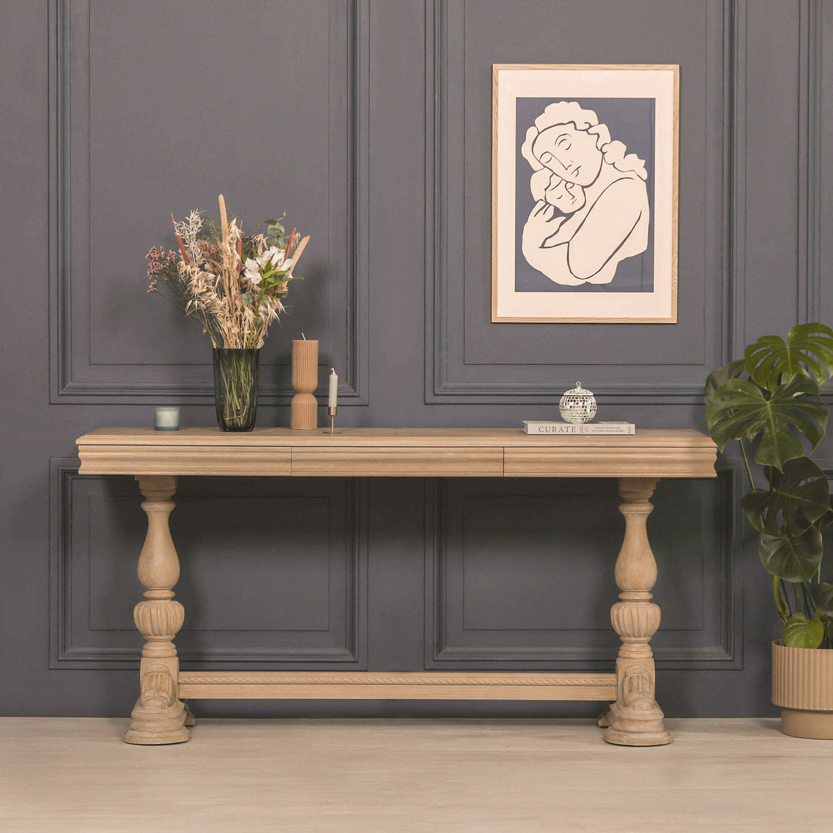 Camille Carved White Cedar Wood Console Table with Drawer in a rustic finish styled against a charcoal panelled wall with decorative accessories, framed artwork and indoor plants, showcasing its timeless French country design and elegant carved craftsmanship.