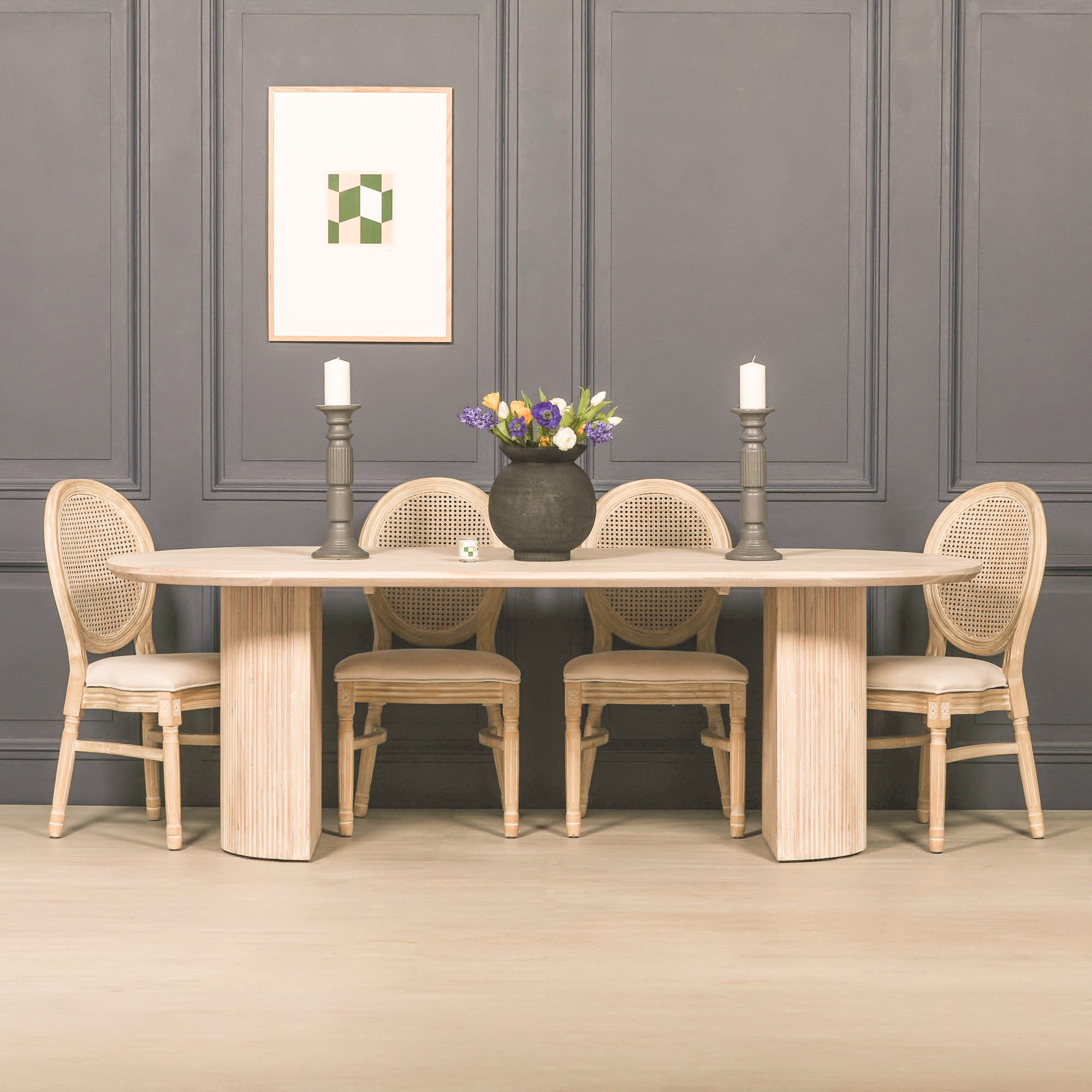 Camille Fluted Acacia Wood Oval 6 Seater Dining Table in a bleached washed wood finish styled with rattan dining chairs in a modern dining room, showcasing the sculptural fluted pedestal legs and spacious oval tabletop for contemporary and coastal interiors.