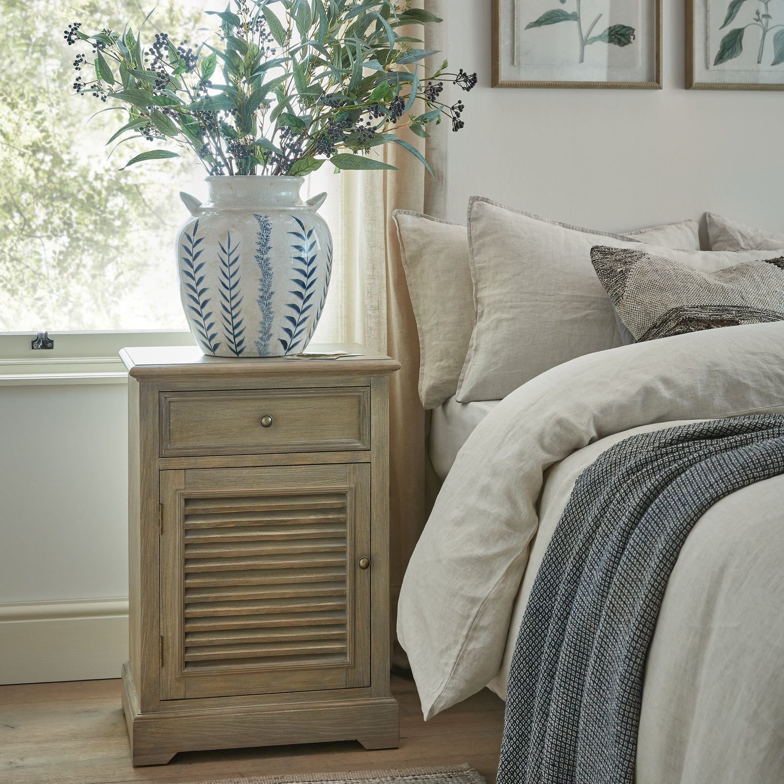 Laurent Wood Storage Chest with 1 Door & 1 Drawer – Left Hand styled as a bedside cabinet in a contemporary bedroom, featuring a natural wood finish, left-hand shutter-style door, spacious storage drawer and practical bedside storage beside a bed and window.