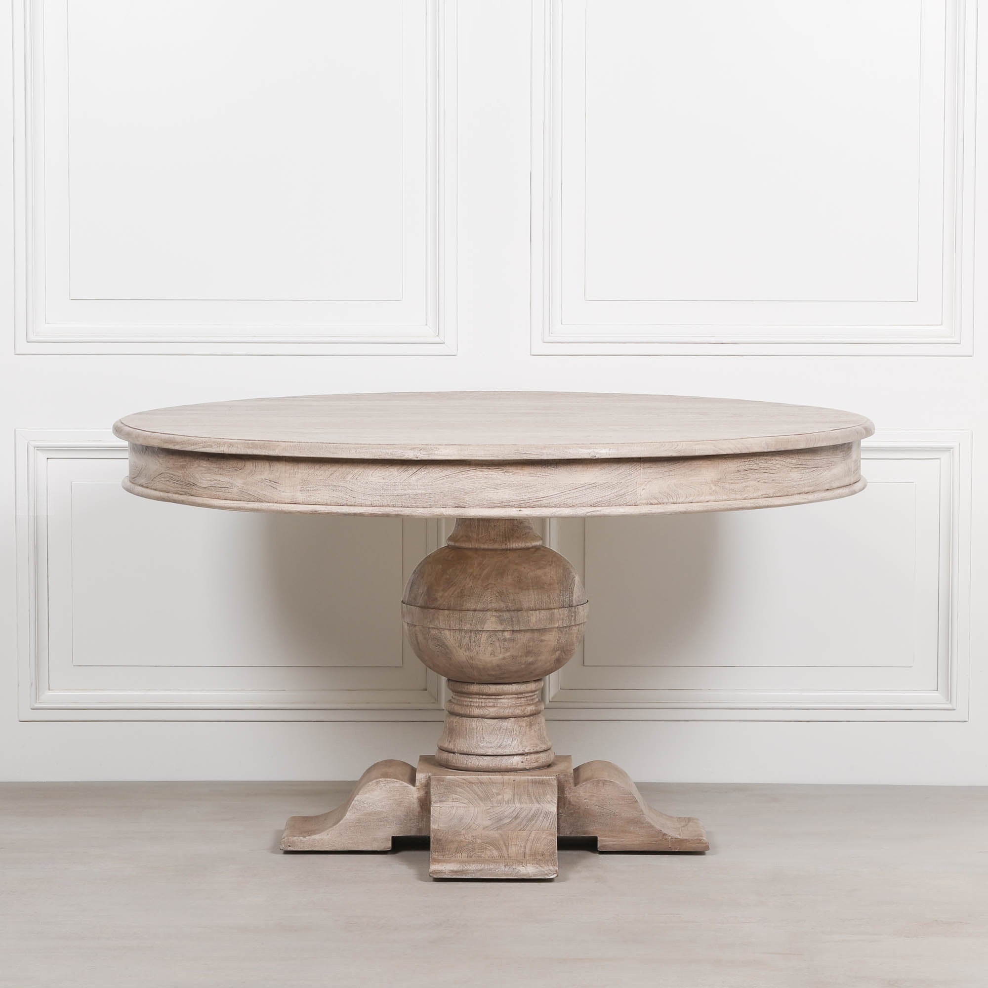 Front view of the Lucan round pedestal dining table in rustic washed solid acacia wood, showcasing the thick circular tabletop, sculptural turned ball pedestal and substantial four-footed base against a classic white panelled backdrop.