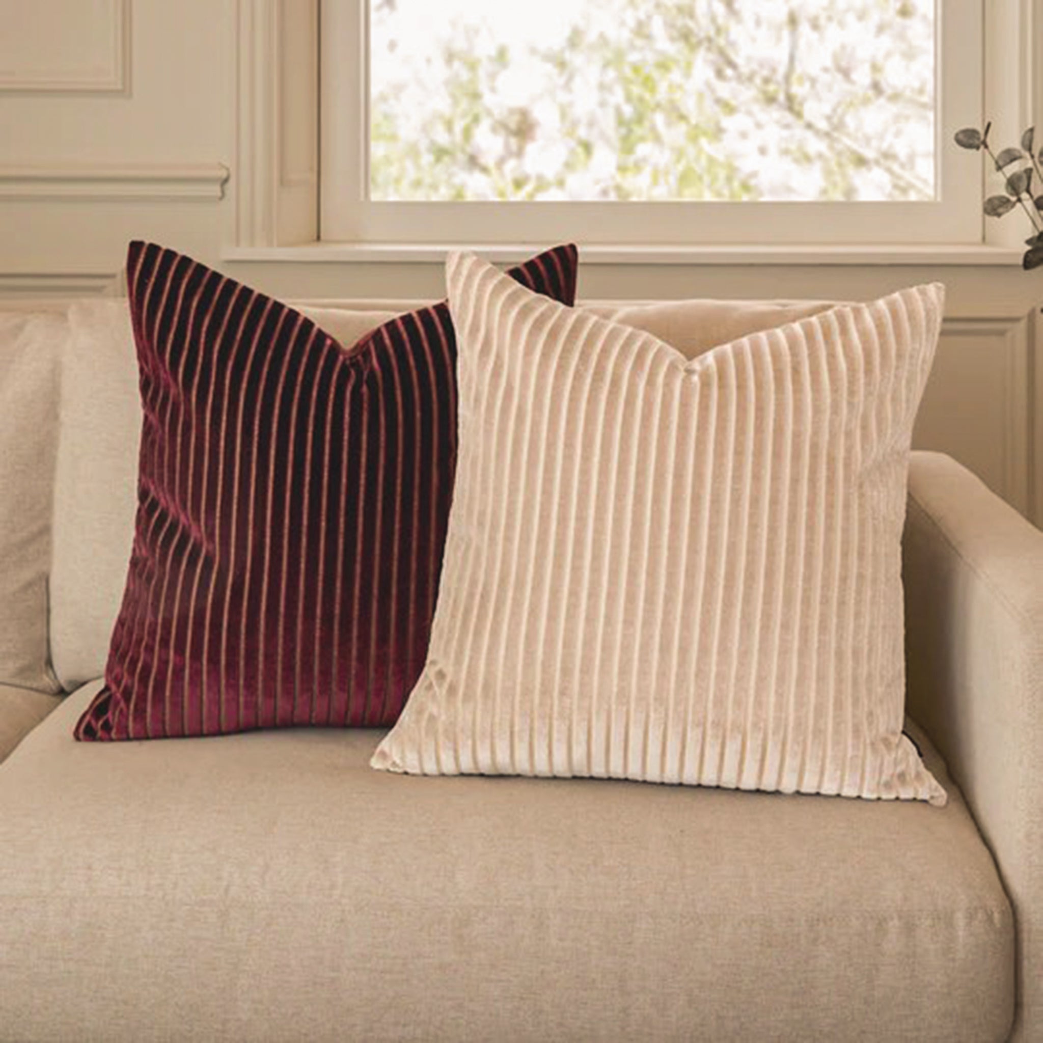 Maëva Cut Velvet Striped Cushion in Oyster Cream styled on a beige sofa alongside a burgundy velvet cushion, showcasing the elegant ribbed cut velvet texture and soft neutral colour palette for luxury living room décor.