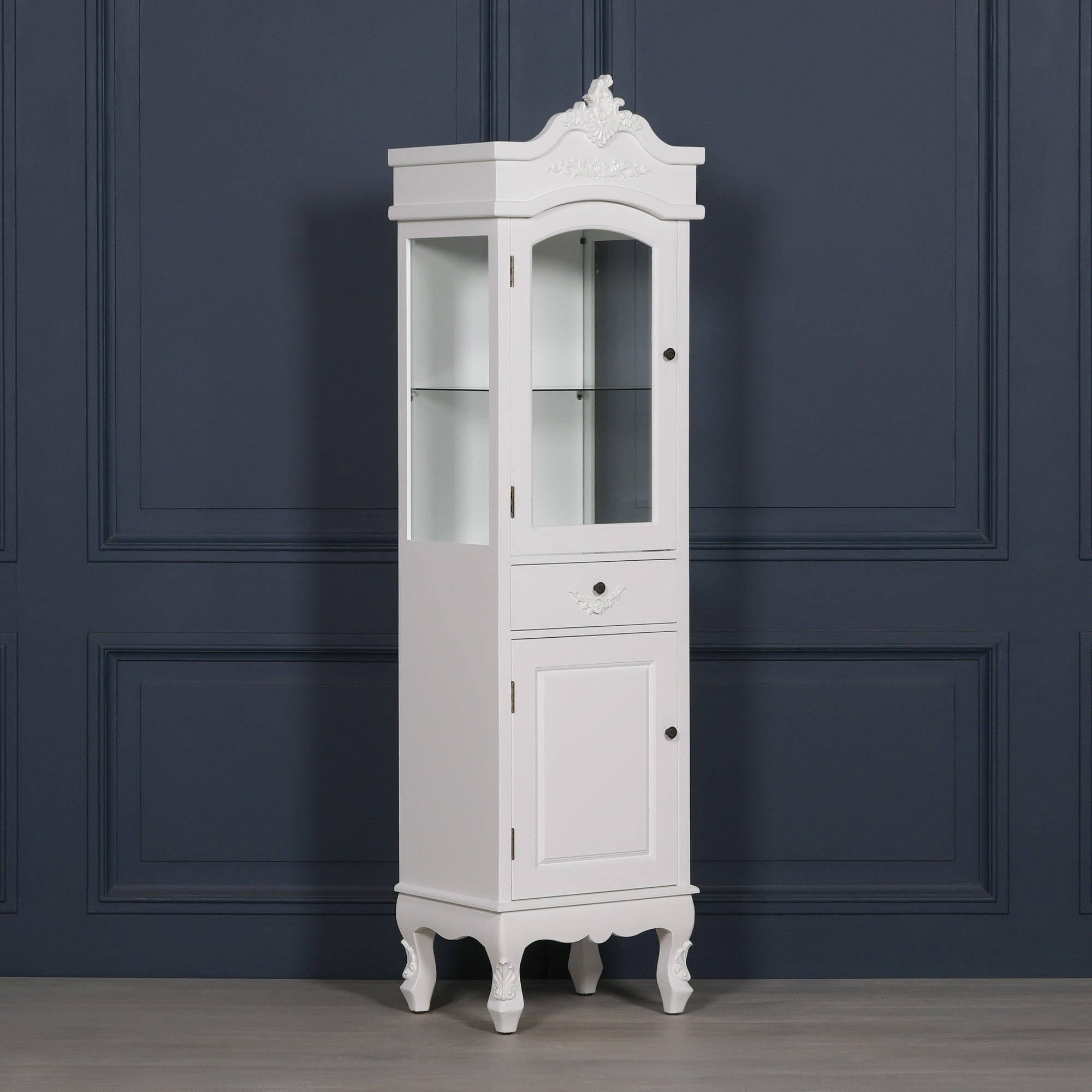 Angled view of the Rosalie Tall French Glass Display Cabinet with Drawer in Antique White featuring glazed display shelves, carved French detailing, decorative cornice, storage drawer and lower cupboard for elegant home interiors.