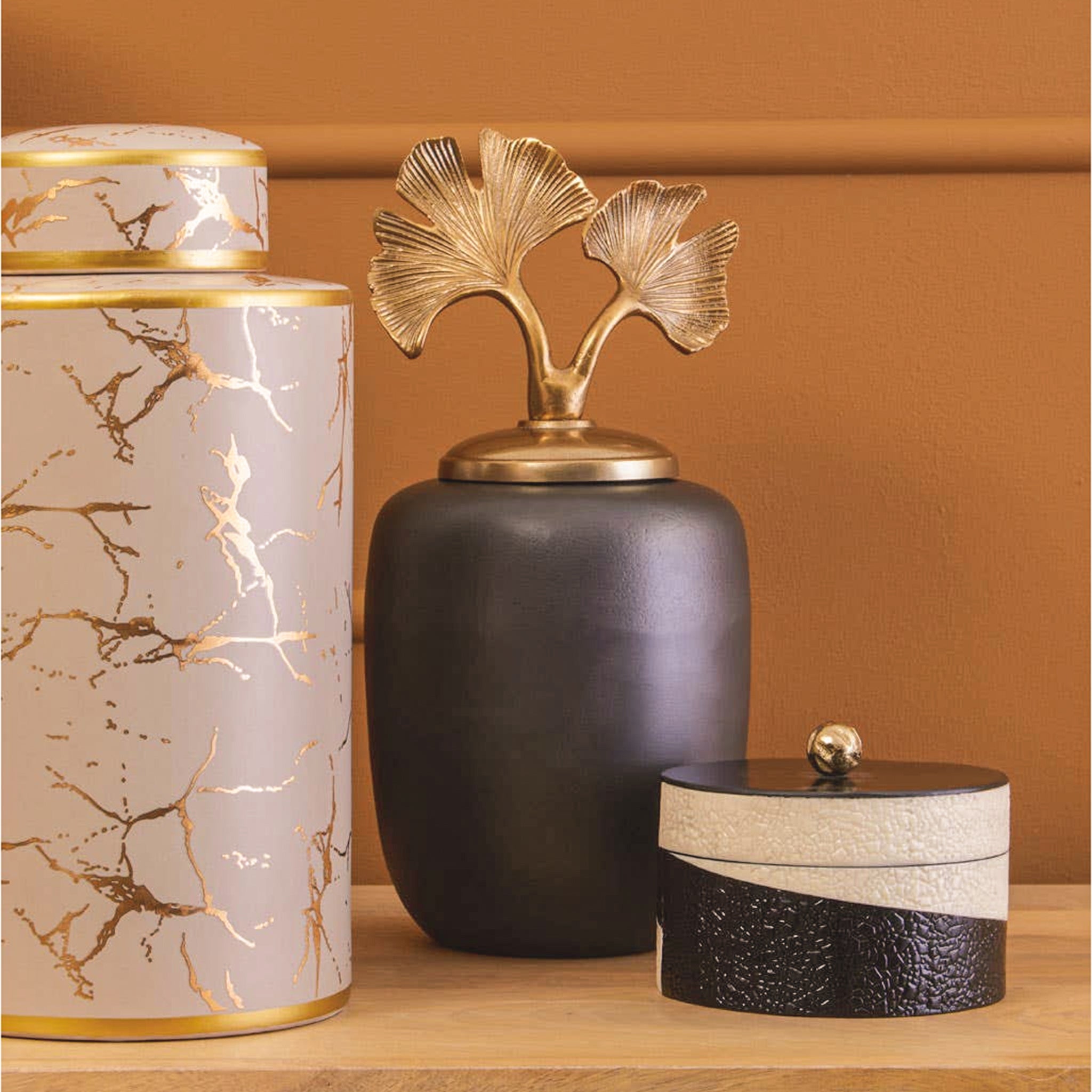 Alix Round Grey Aluminium Ginkgo Leaf Decorative Ginger Jar styled on a wooden surface, featuring a dark grey rounded body and sculptural gold ginkgo leaf lid alongside decorative accessories.