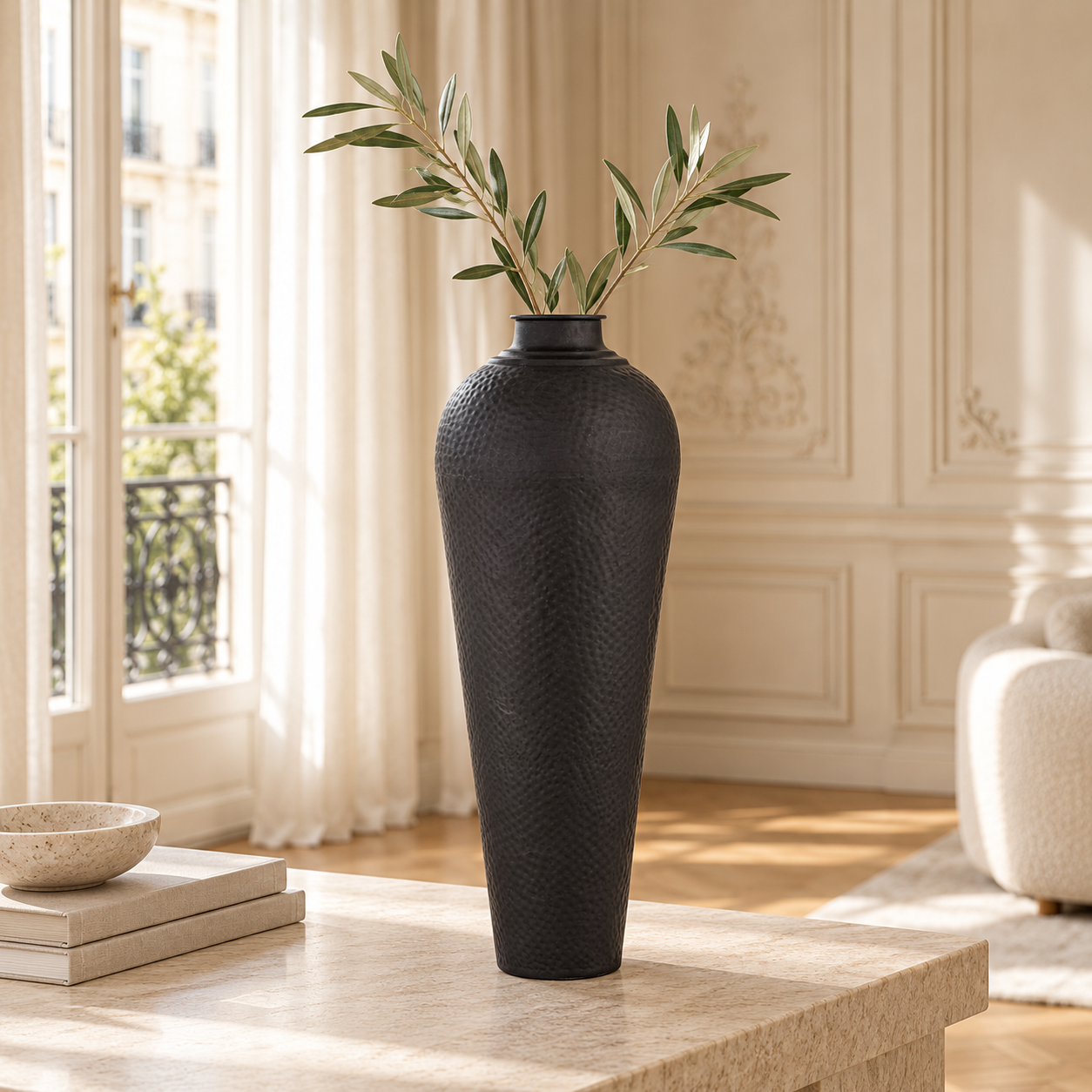 Alix tall hammered black metal vase with olive branches on a stone table in a bright cream living room.