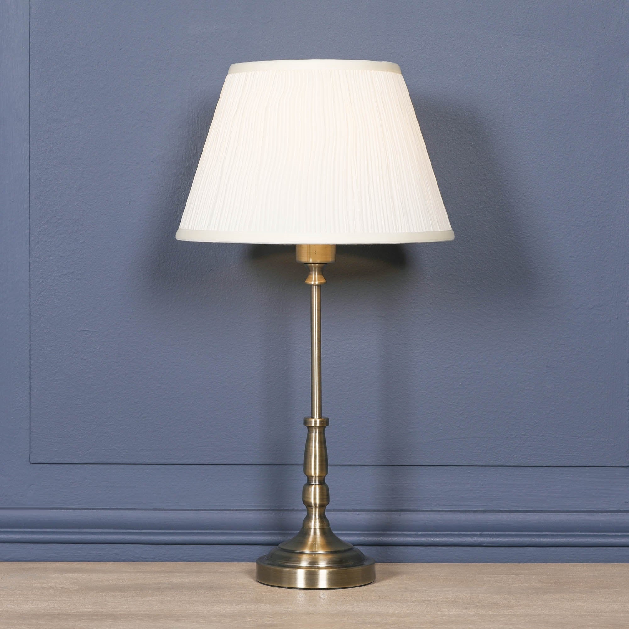 Full view of the Amélie Classical satin brass candlestick table lamp with cream pleated shade on a wooden surface against a blue panelled wall, timeless traditional lighting for luxury interiors.