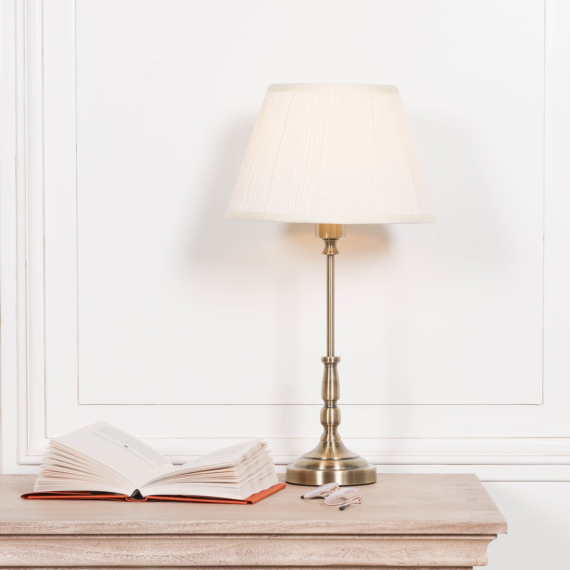 Amélie Classical satin brass candlestick table lamp with cream pleated shade styled on a wooden console table beside an open book and reading glasses, traditional luxury bedside lighting for elegant living rooms and bedrooms.