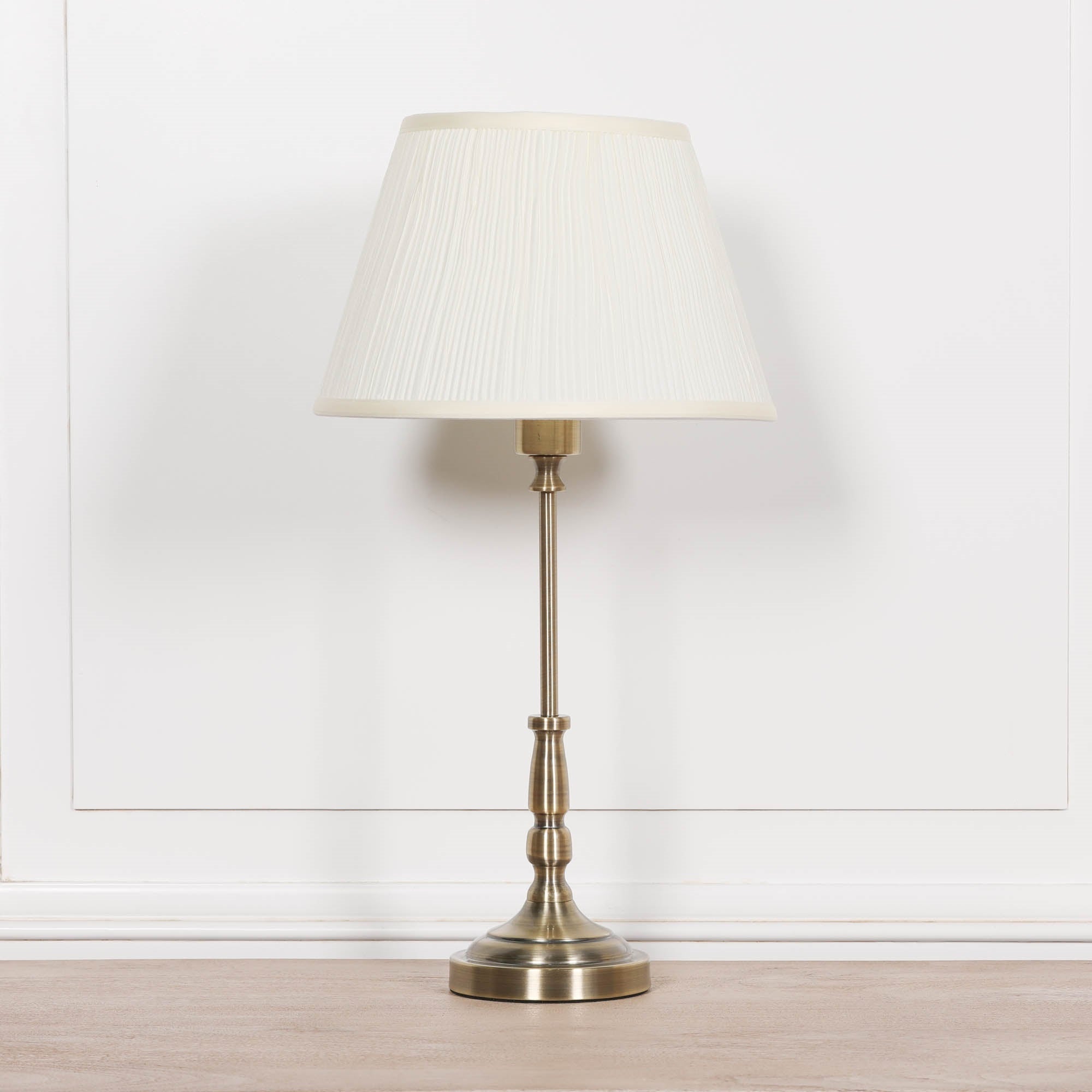 Front view of the Amélie Classical satin brass candlestick table lamp with cream pleated shade displayed on a wooden tabletop against a white panelled wall, ideal as a bedside or living room table lamp.