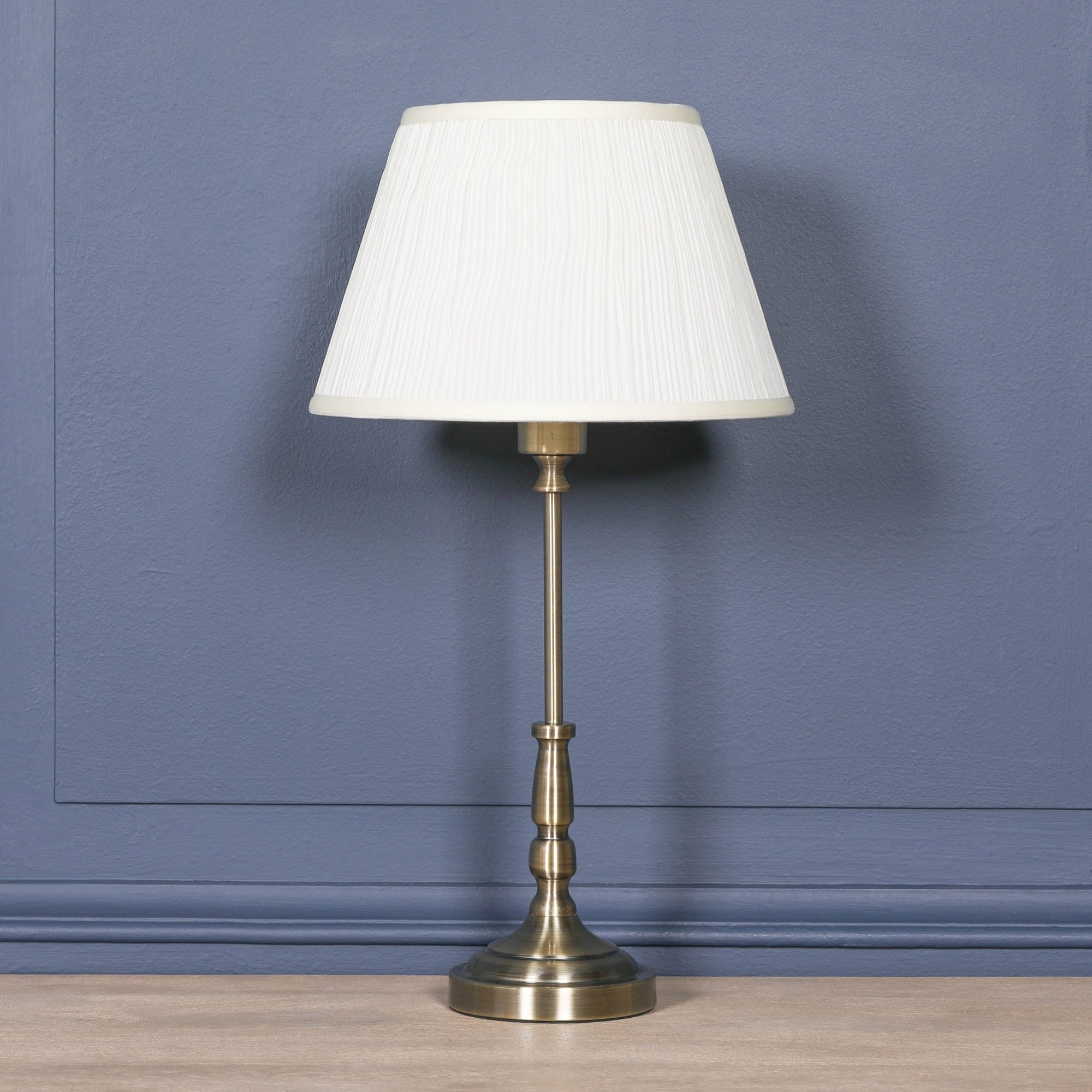 Amélie Classical satin brass candlestick table lamp with cream pleated shade displayed against a blue panelled wall, perfect as a traditional bedside lamp or elegant living room table lamp.