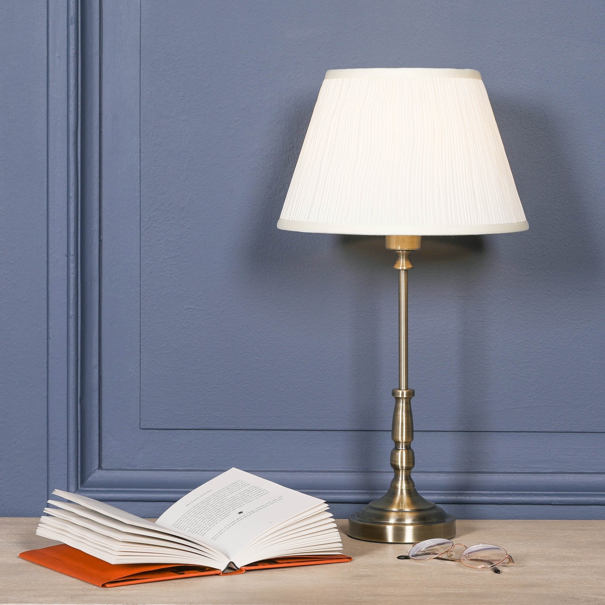Amélie Classical satin brass candlestick table lamp with cream pleated shade styled beside an open book and reading glasses on a console table against a blue wall, creating a sophisticated reading nook.