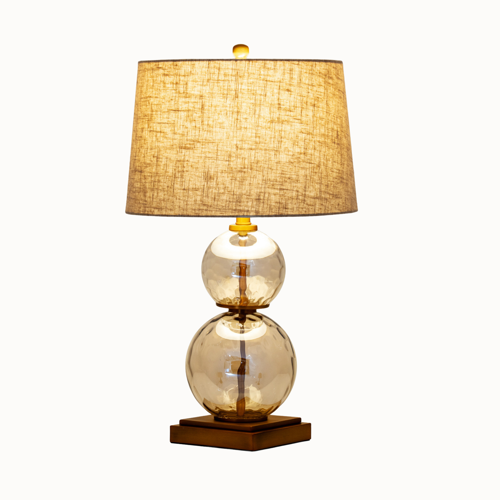 Clear glass and gold table lamp illuminated through its linen shade against a plain white background.