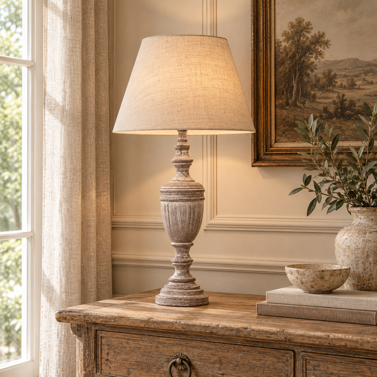 Amélie grey wooden urn table lamp with linen shade on a rustic console in a bright classic room with window and artwork.