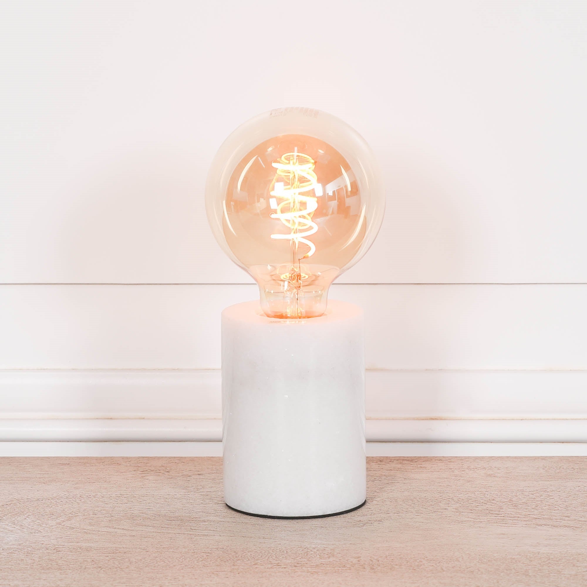 Amélie industrial white marble table lamp with exposed glowing bulb on a wood surface against a white wall.