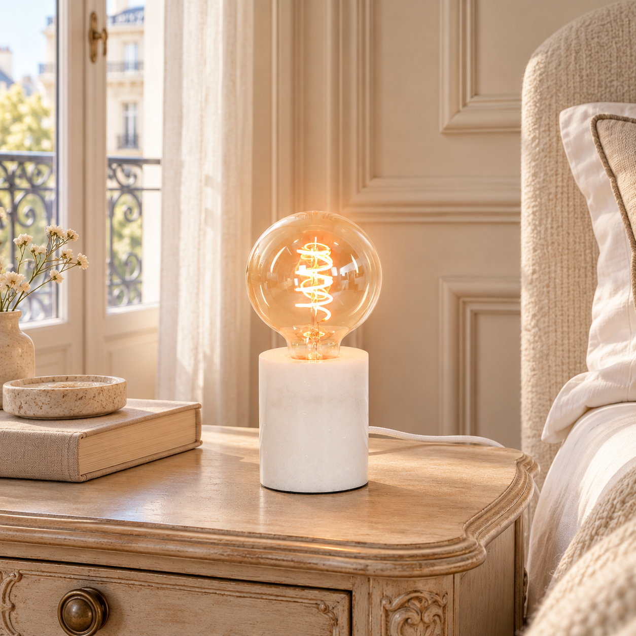 Amélie industrial white marble table lamp with exposed bulb on a bedside table in a sunlit Parisian bedroom.
