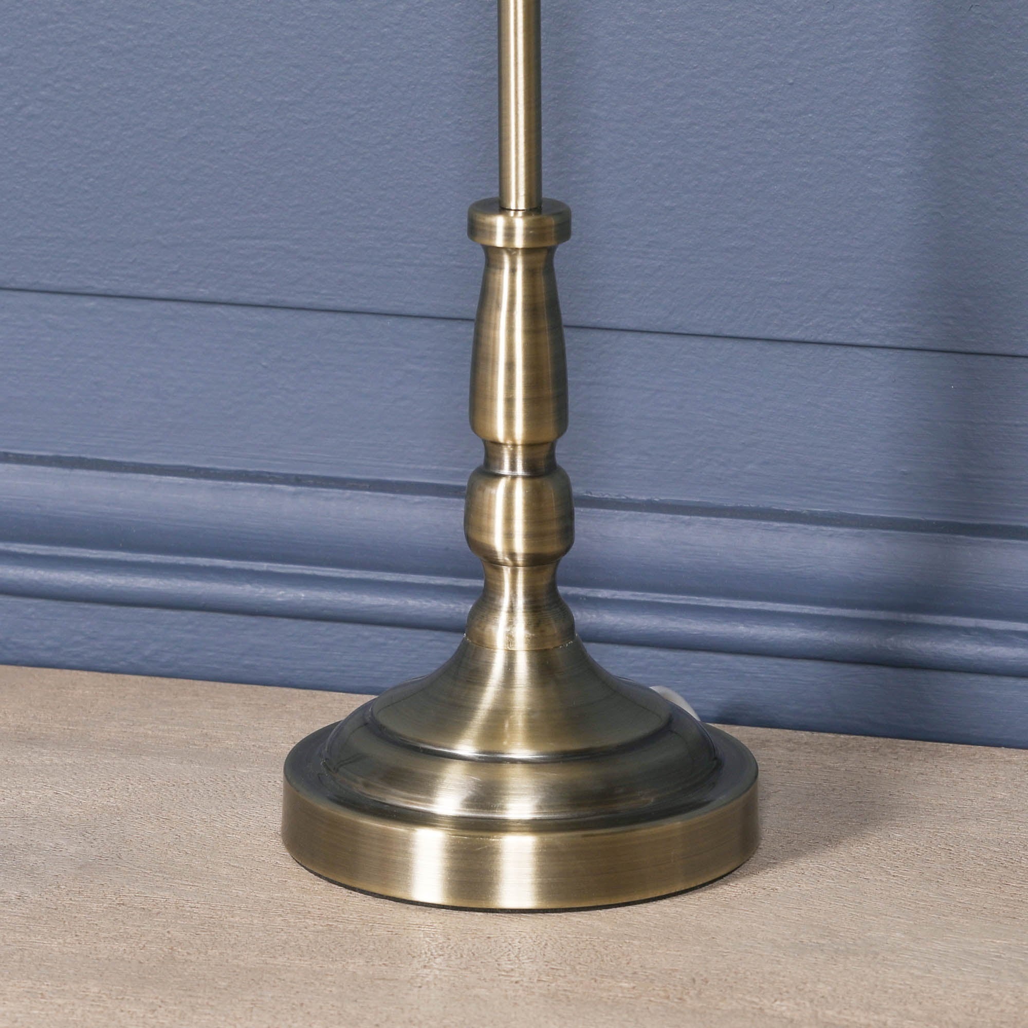Detailed view of the satin brass candlestick base of the Amélie Classical table lamp on a wooden tabletop against a blue panelled wall, highlighting the premium brushed brass finish.