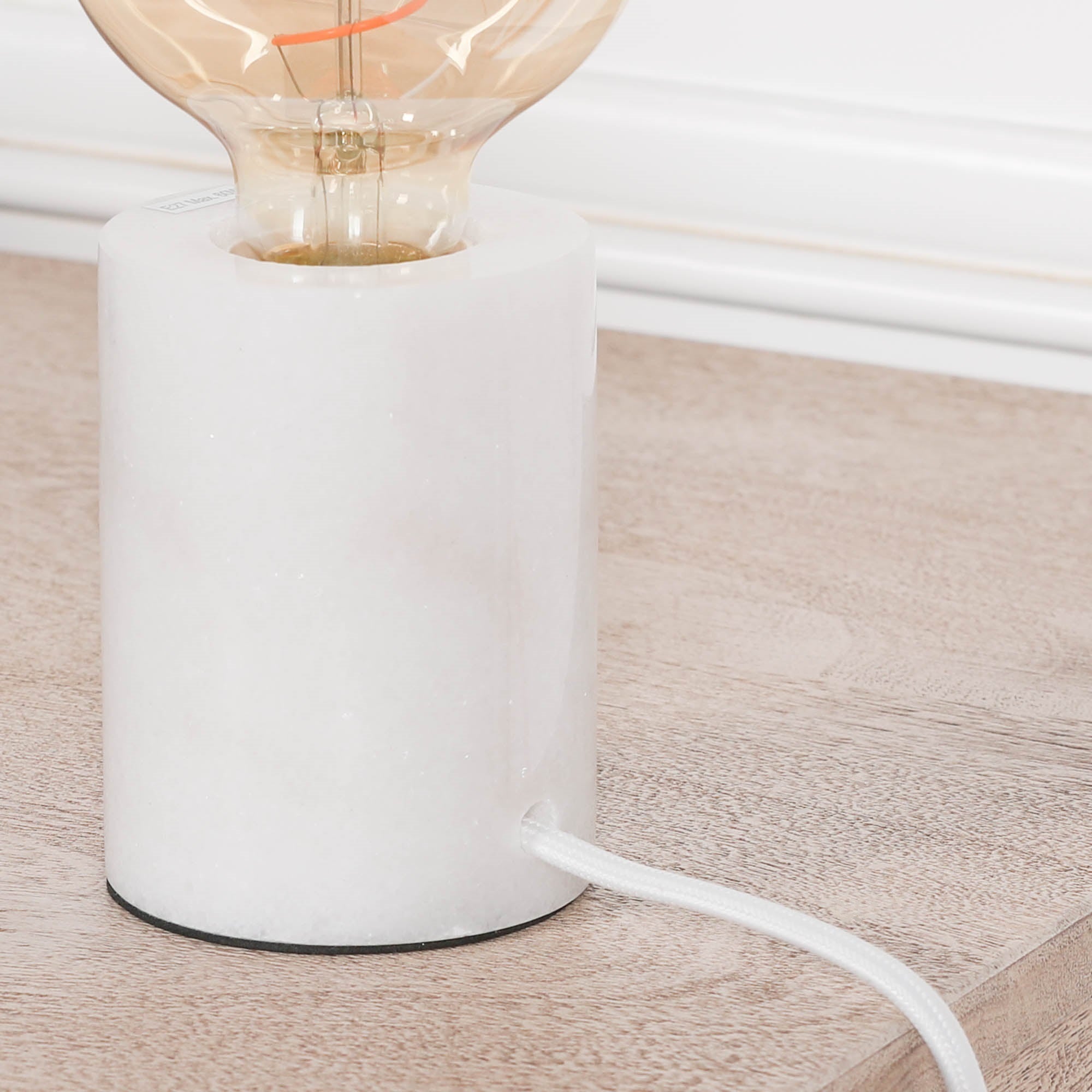 Close-up of Amélie white marble table lamp showing cylindrical base, exposed bulb and braided cord on a wood surface.
