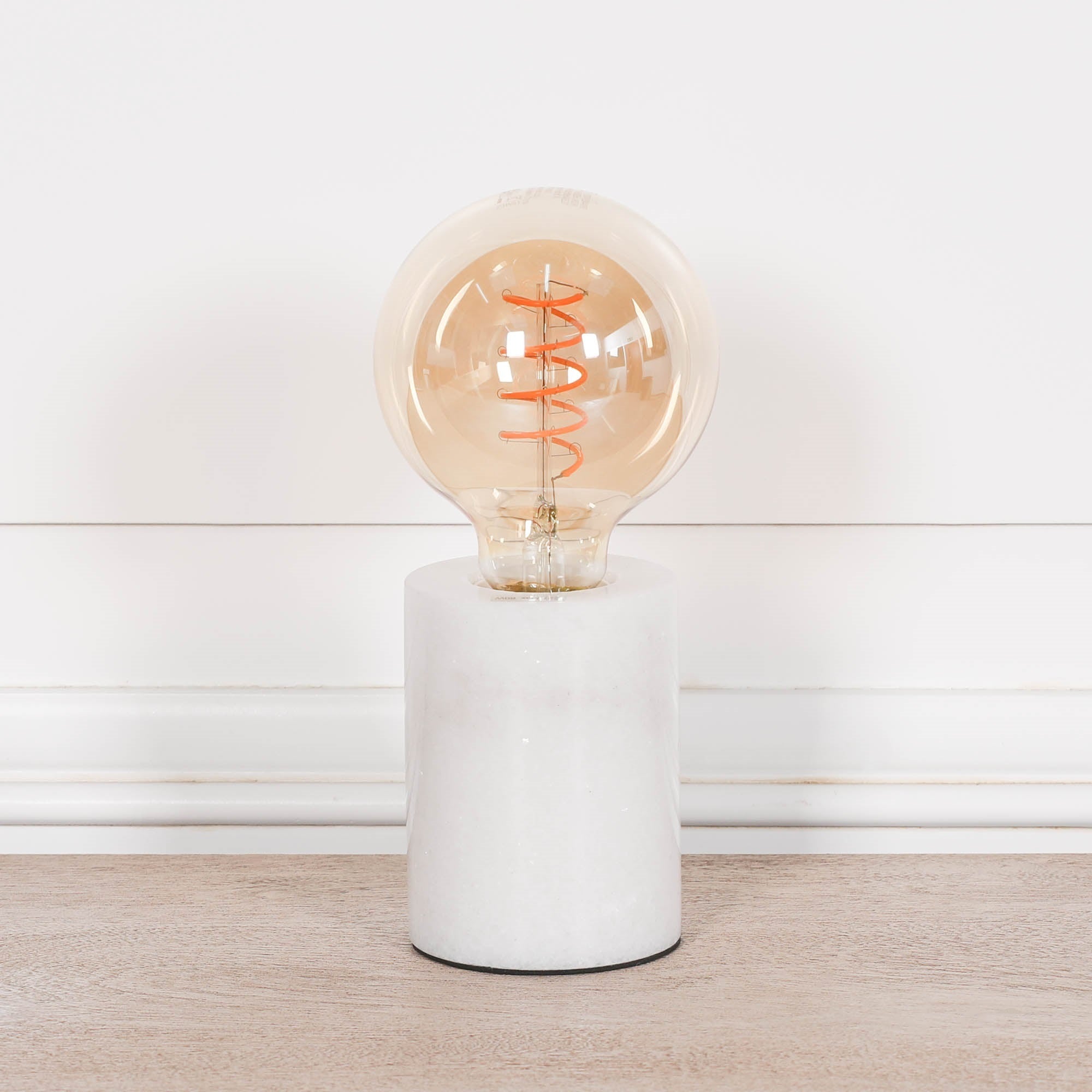 Amélie white marble table lamp with exposed bulb on a light wood surface against a clean white panelled wall.