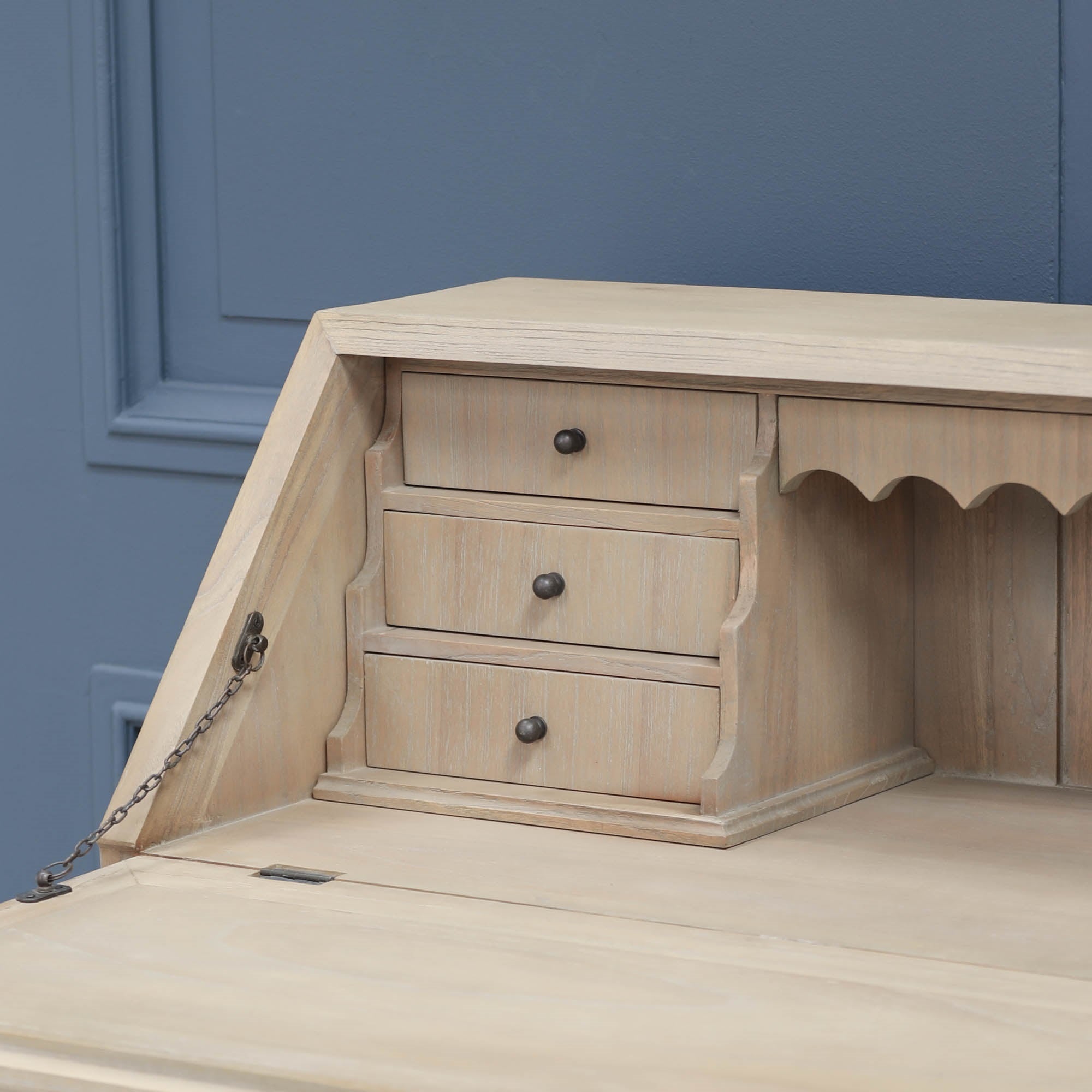 Armand white cedar bureau desk close-up showing three drawers and drop-front chain against a blue panelled wall.