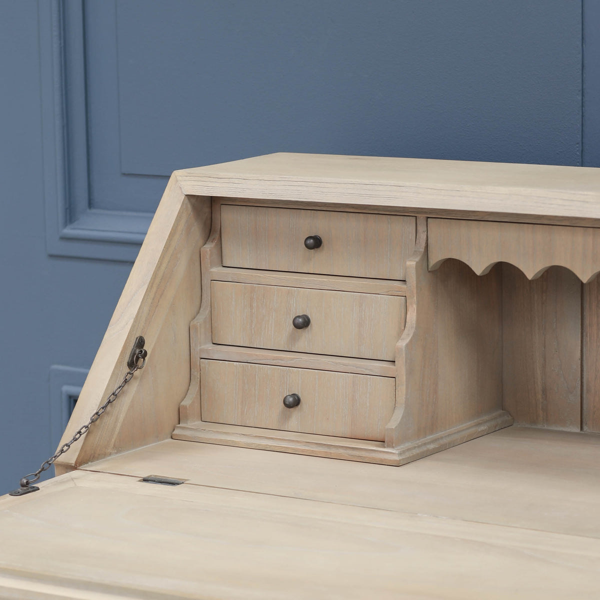 Armand white cedar bureau desk close-up showing three drawers and drop-front chain against a blue panelled wall.