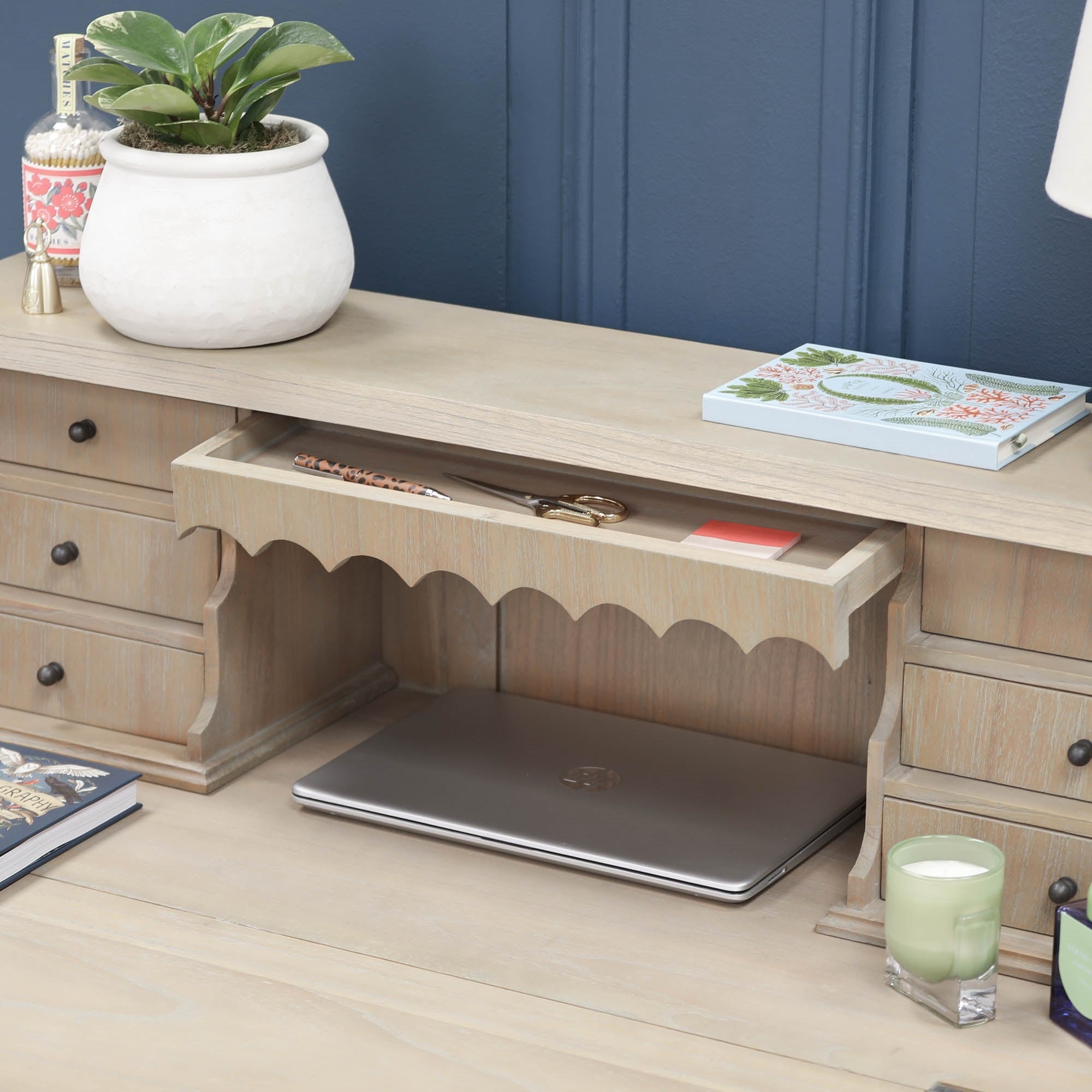Armand white cedar bureau desk close-up with open centre drawer, laptop and plant against a blue panelled wall.