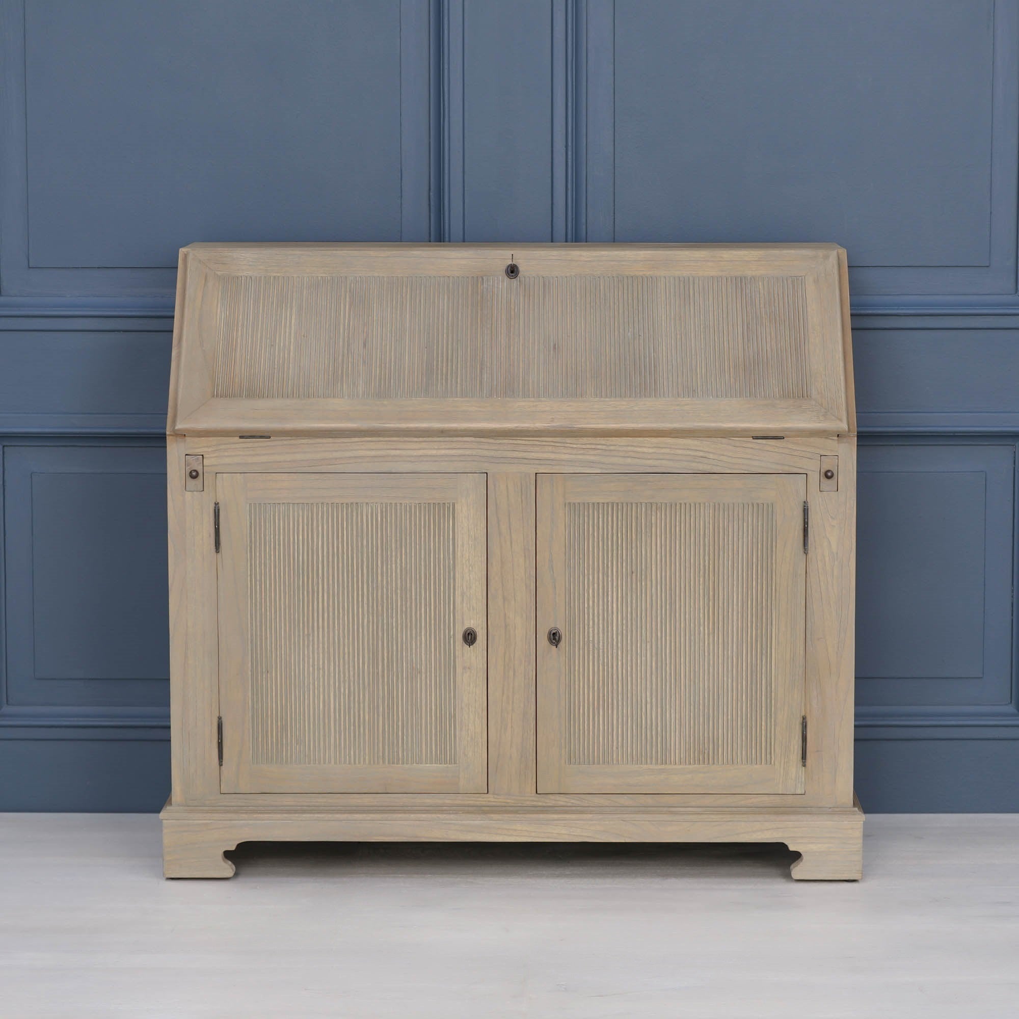 Armand white cedar bureau writing desk closed with reeded storage doors against a blue panelled wall.