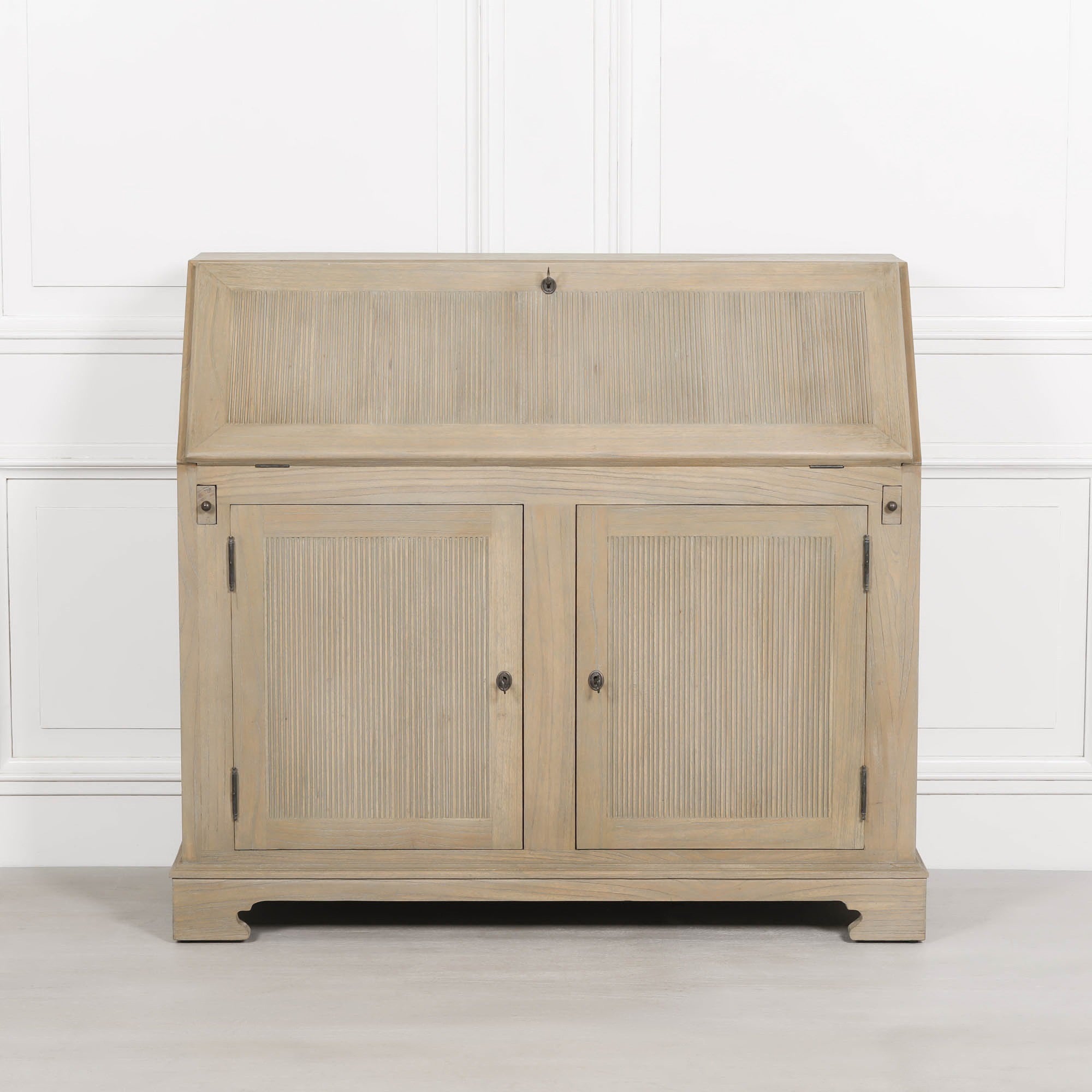 Armand white cedar bureau writing desk closed with reeded storage doors against a white panelled wall.