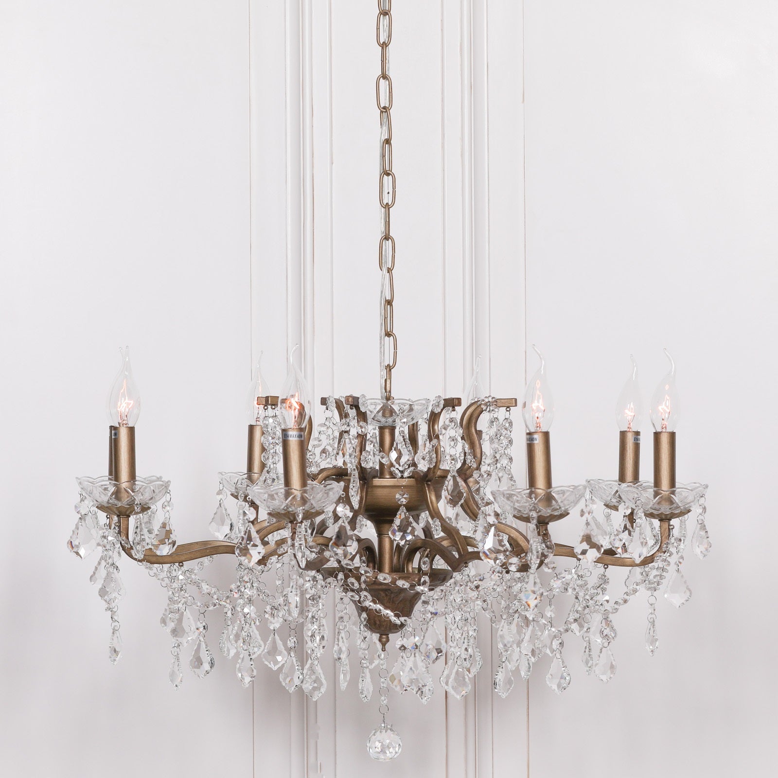 Gold metal 8 light chandelier with clear cut glass drops and candle bulbs against a white panelled wall.