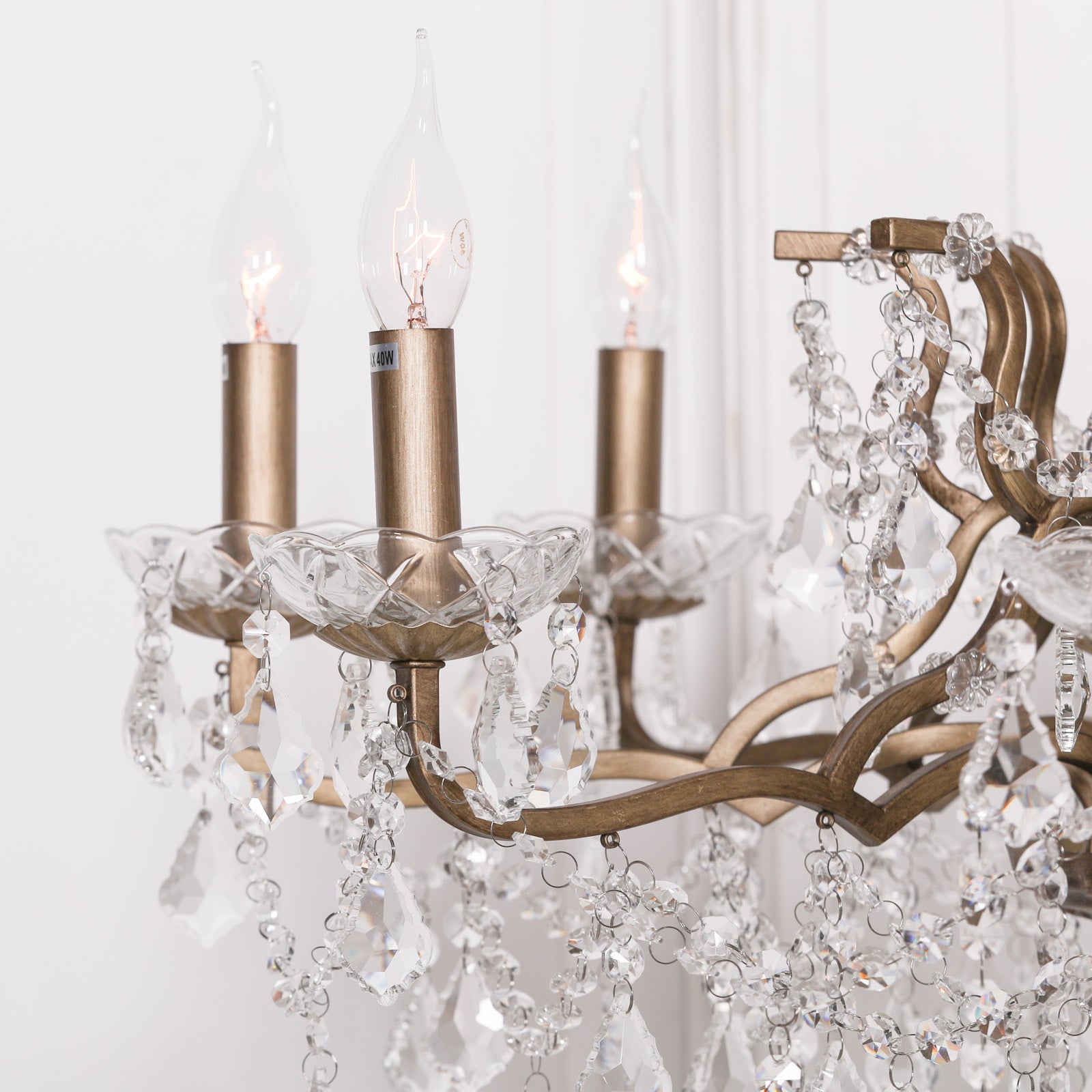 Close-up of Aubert gold chandelier arms, candle bulbs and clear cut glass drops against a white wall.