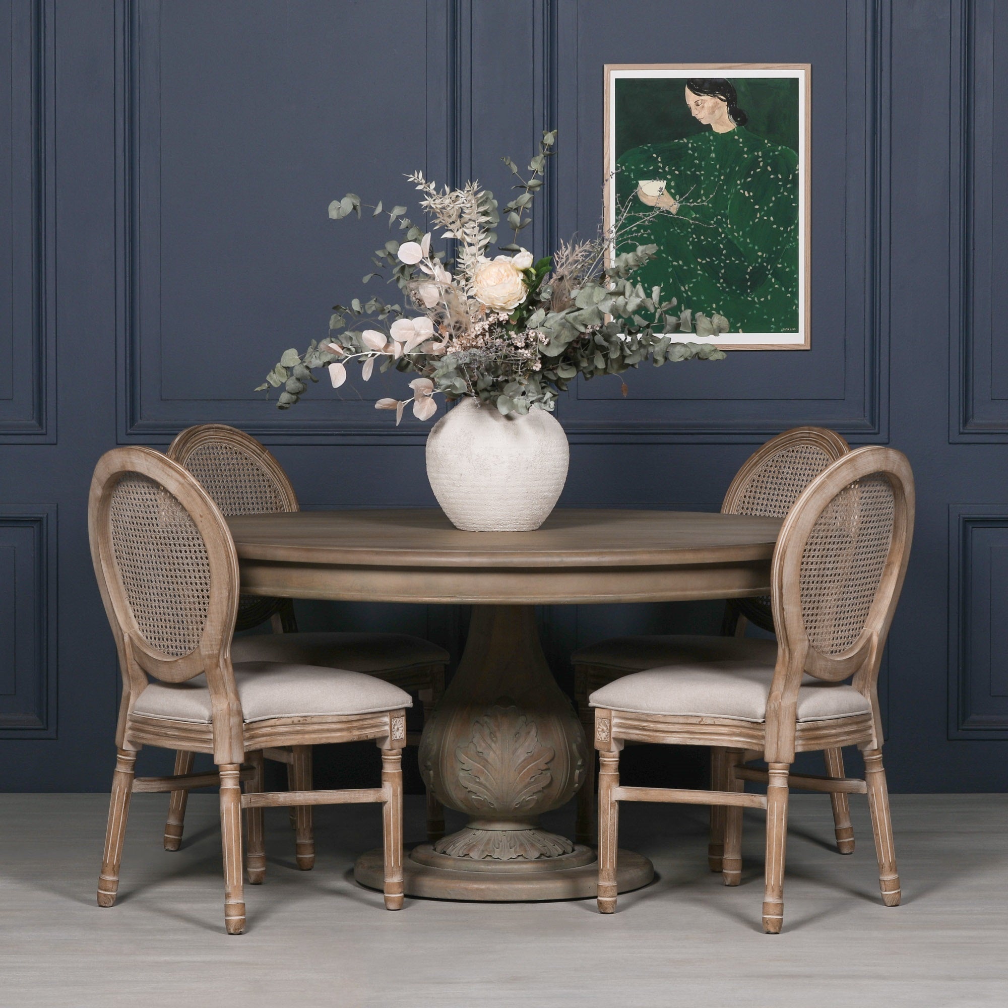 Auguste Round Acorn Pedestal Dining Table 150cm for 6 in Rustic Washed Hardwood styled with upholstered rattan dining chairs, elegant floral arrangement and decorative artwork, showcasing the handcrafted round tabletop, carved acorn pedestal base and luxury farmhouse dining furniture for six people.