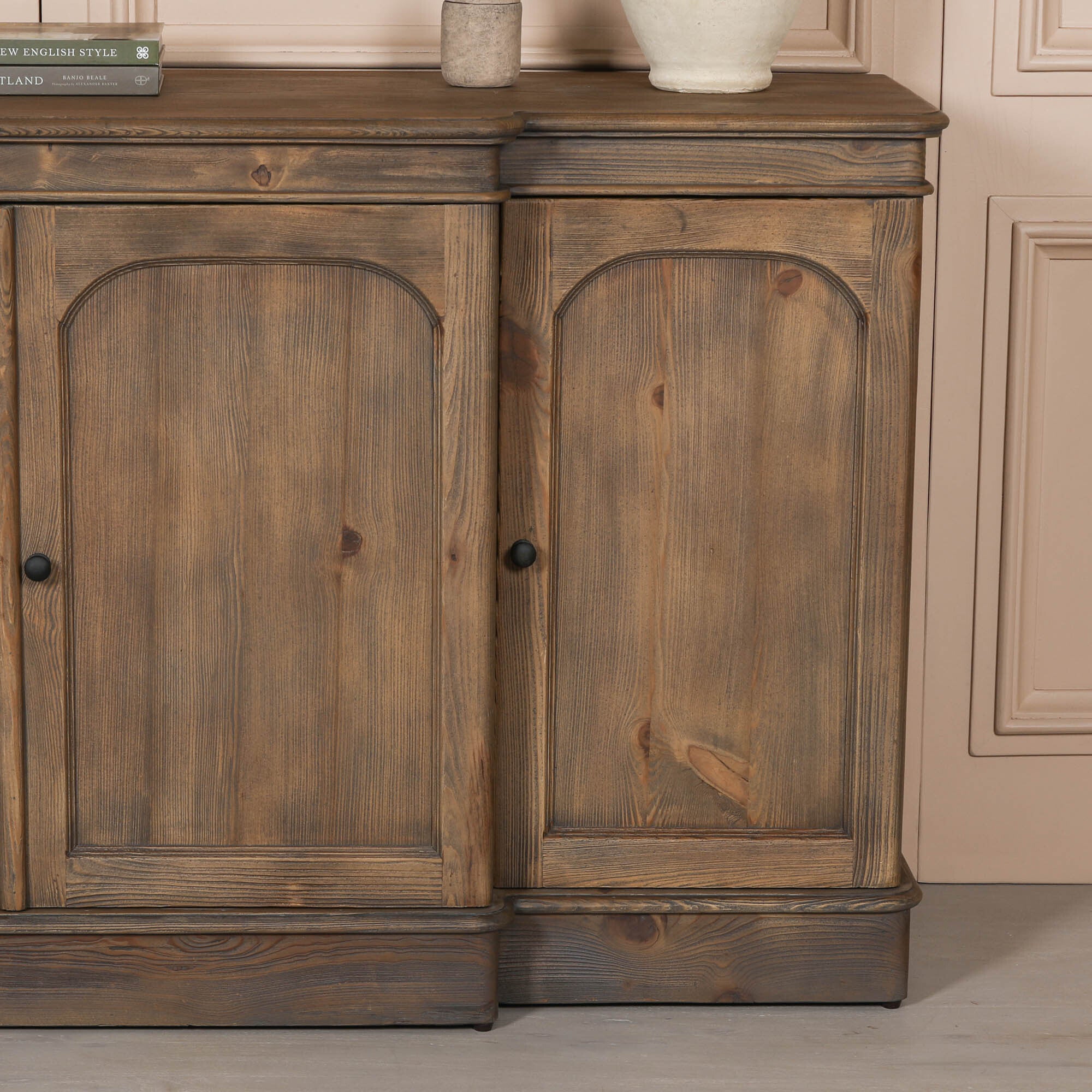 Close-up of Aveline rustic pine wood sideboard with arched doors and black knobs against a neutral panelled wall.