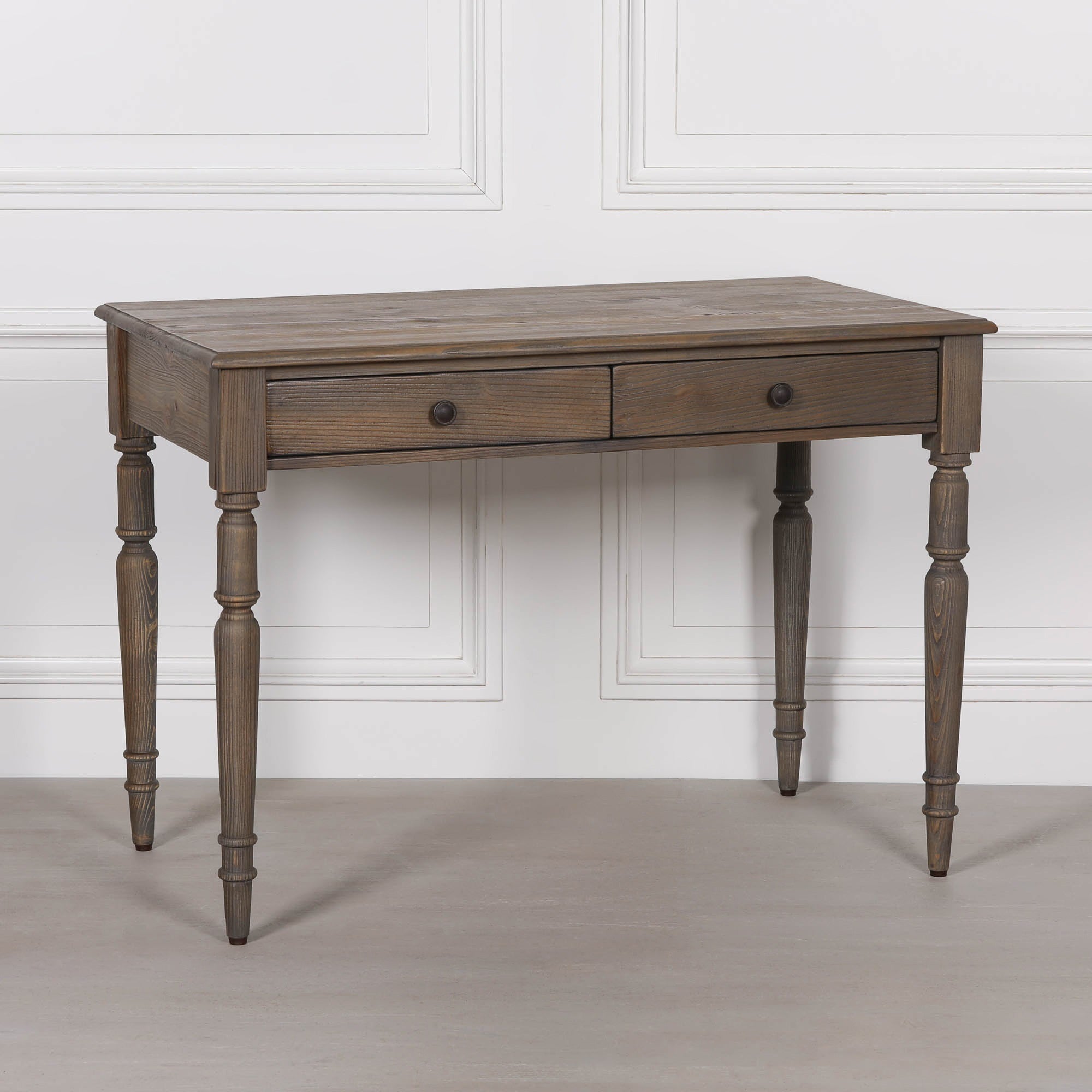 Aveline pine wood writing desk with 2 drawers and turned legs against a white panelled wall and neutral floor