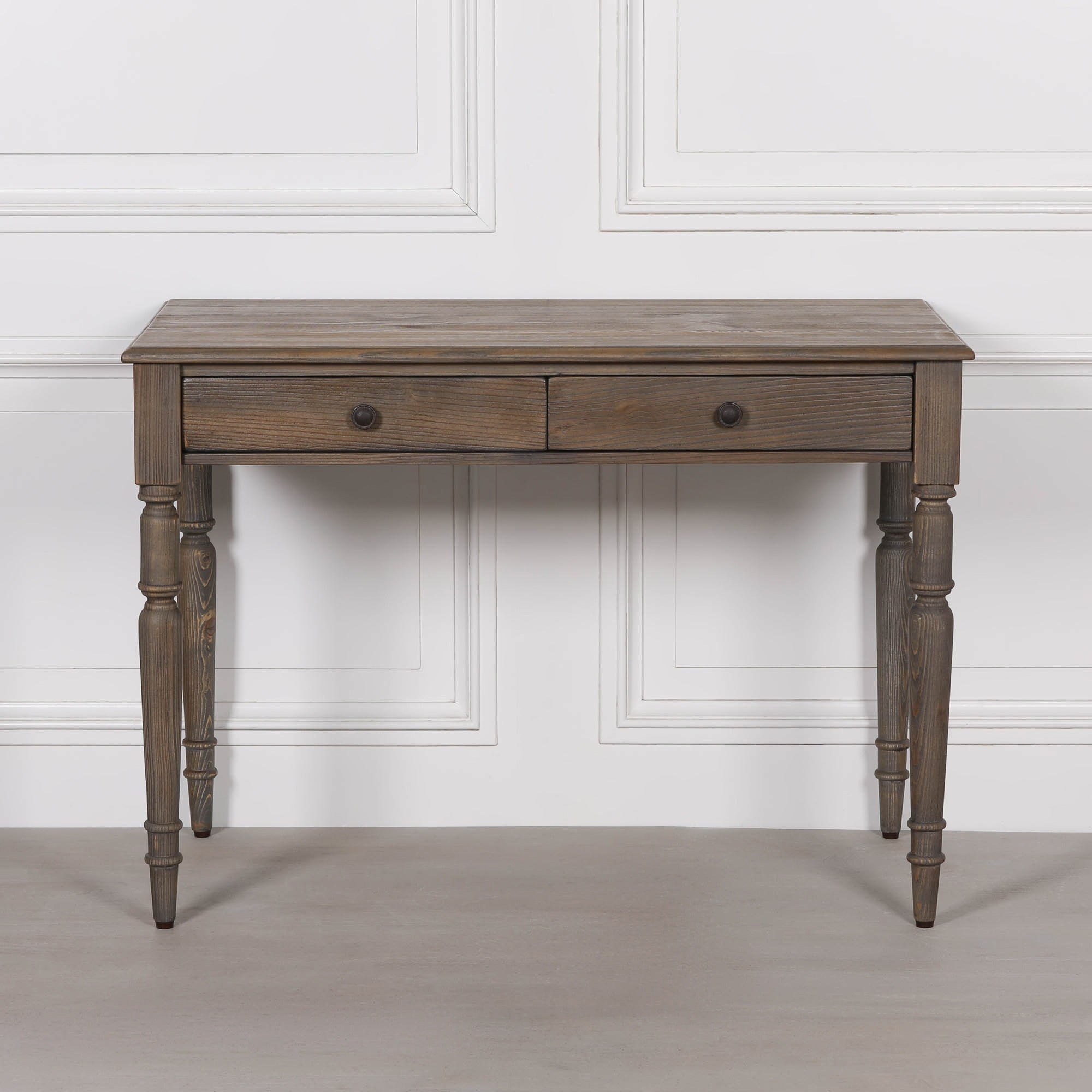 Aveline pine wood writing desk with 2 drawers and turned legs against a white panelled wall and grey floor