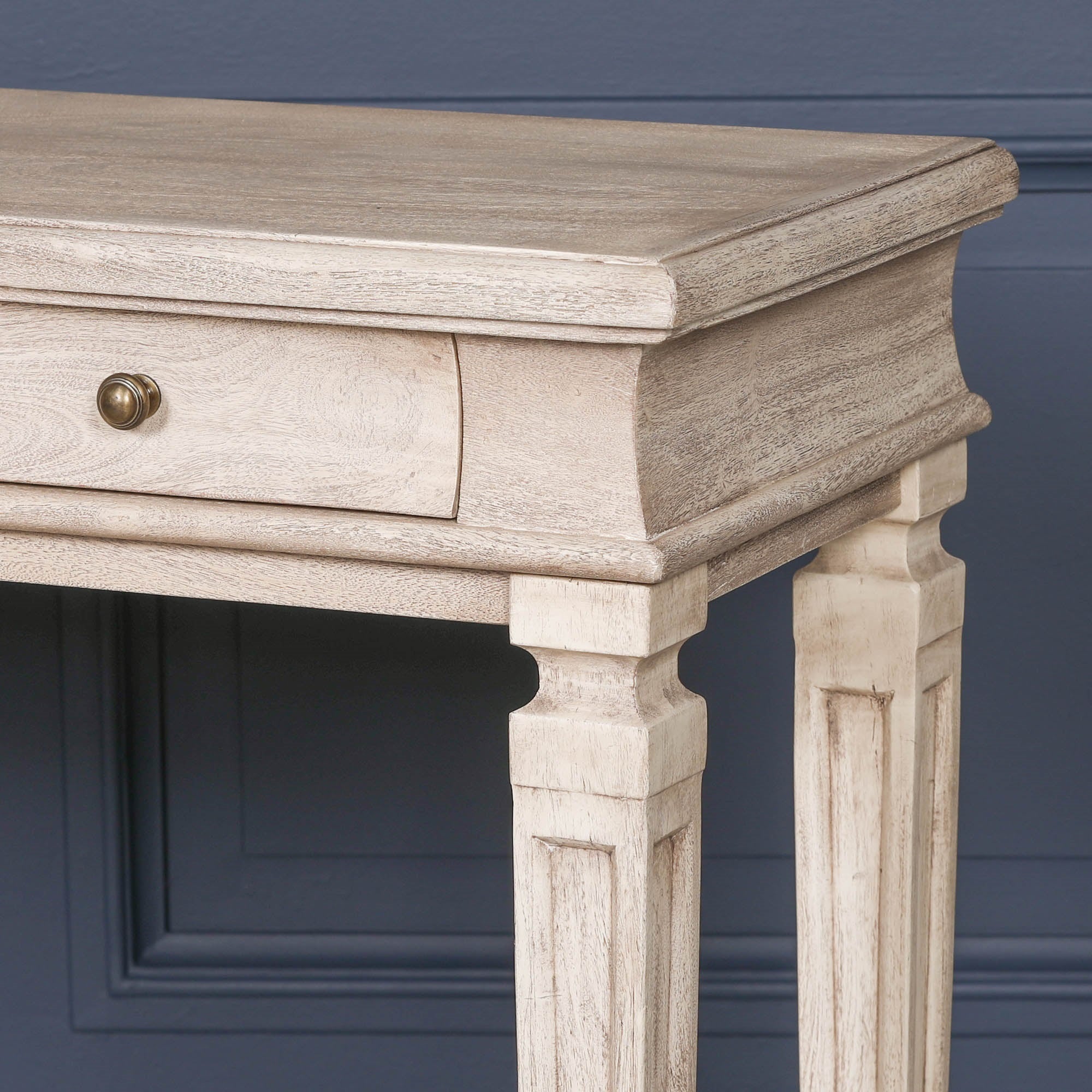 Close-up of Camille acacia wood breakfront console table drawer and carved leg against a dark blue panelled wall