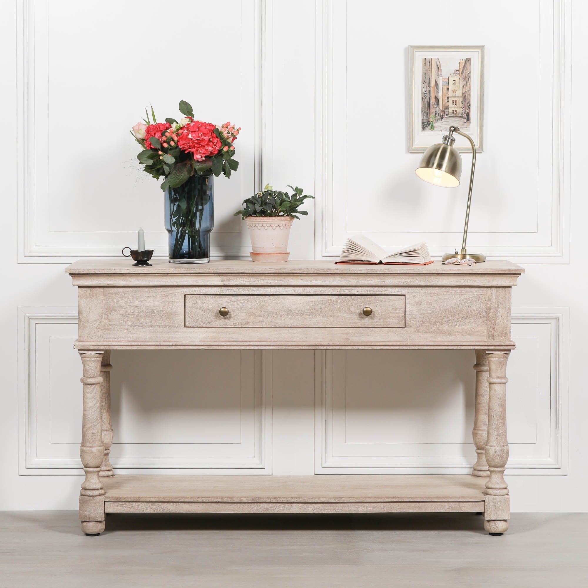 Camille acacia wood console table with drawer and lower shelf styled with flowers and lamp against a white panelled wall