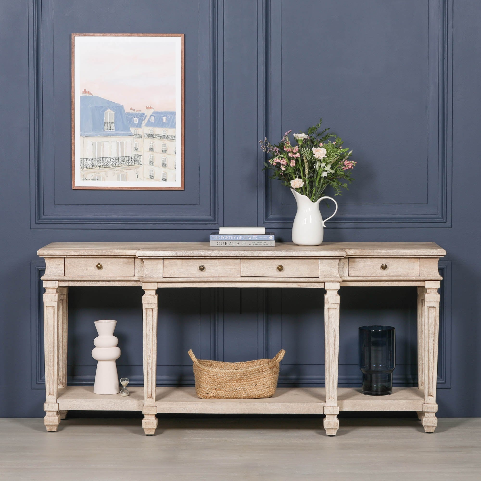 Camille acacia wood 4 drawer breakfront console table styled with flowers against a dark blue panelled wall
