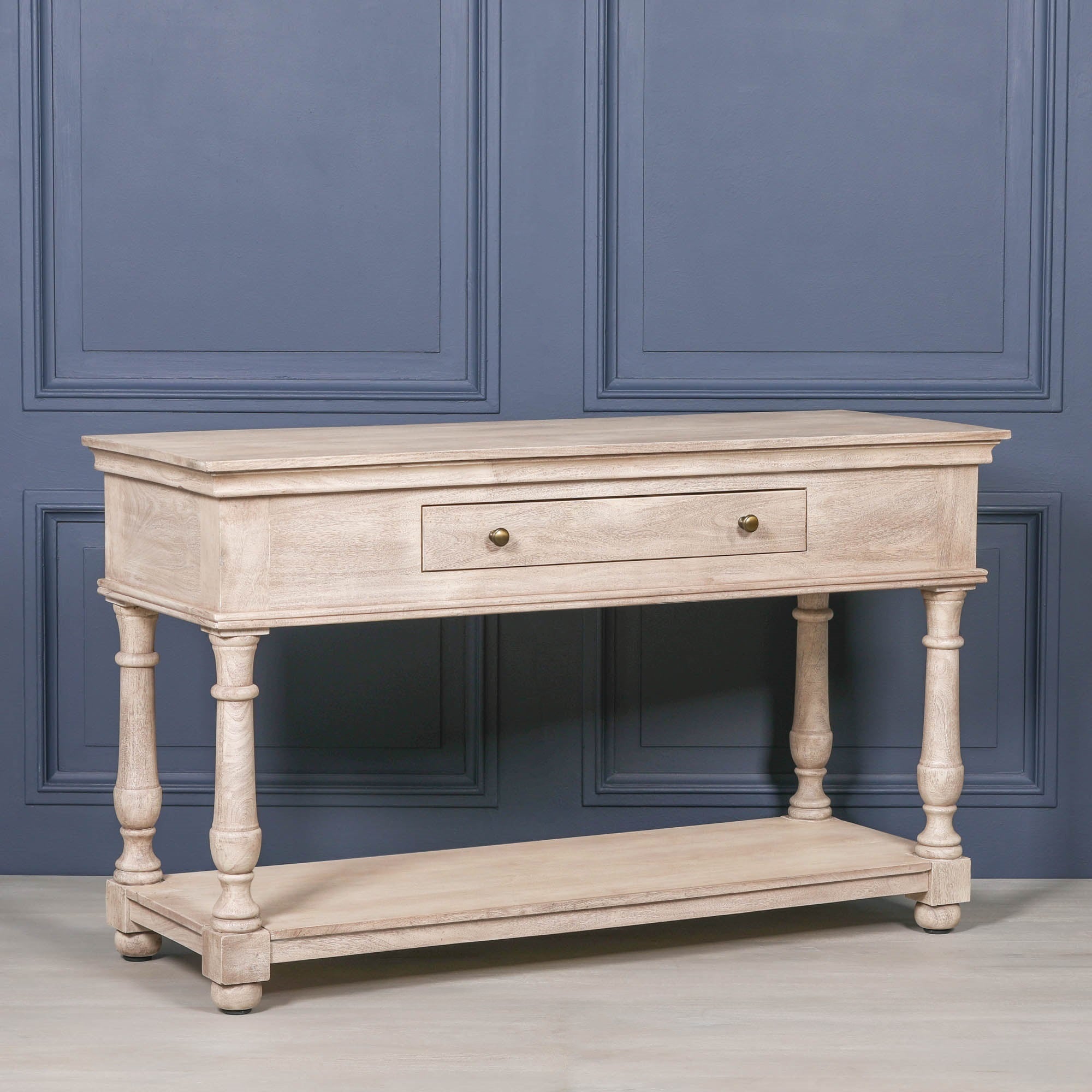 Angled view of Camille acacia wood console table with drawer and lower shelf against a dark blue panelled wall