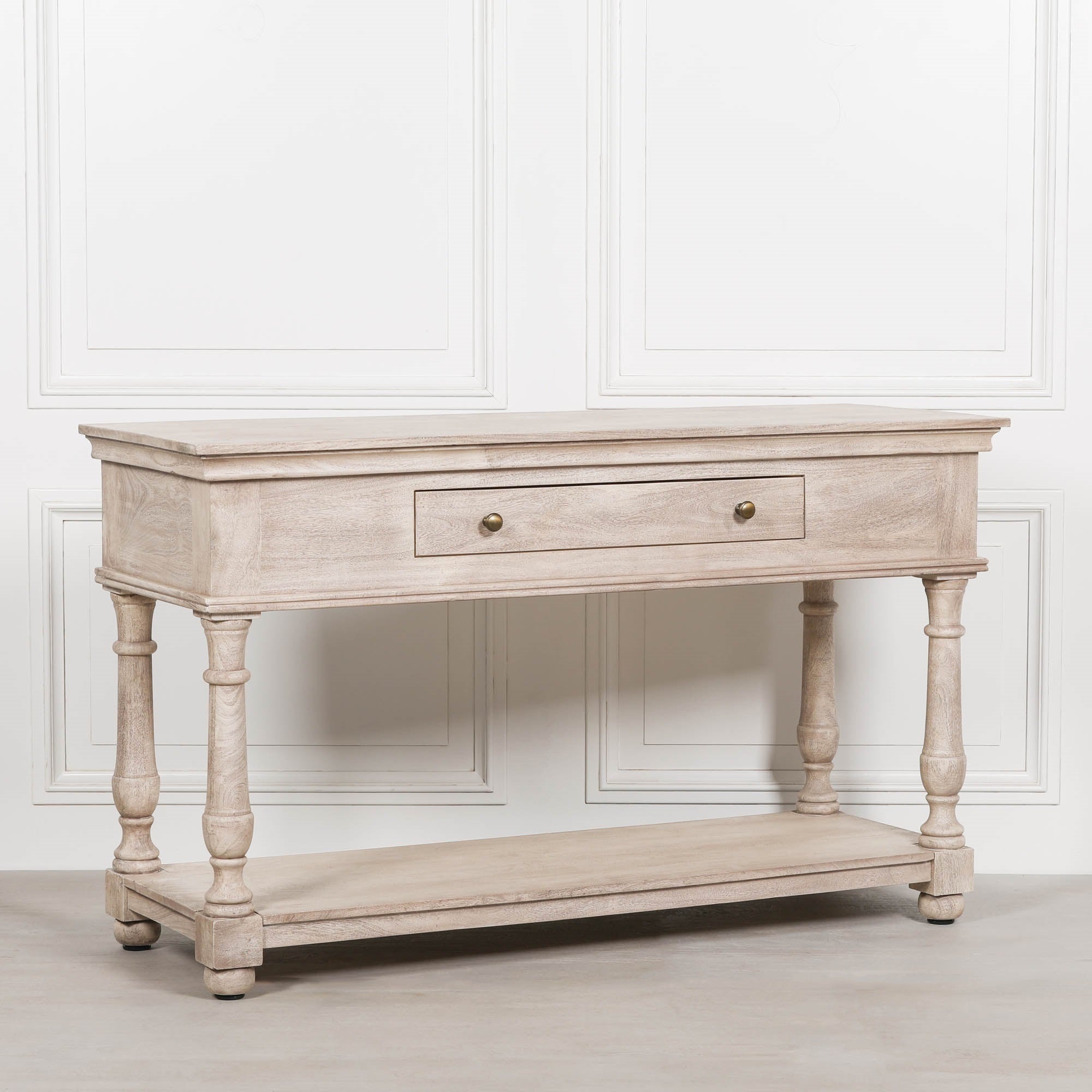 Angled view of Camille acacia wood console table with drawer and lower shelf against a white panelled wall