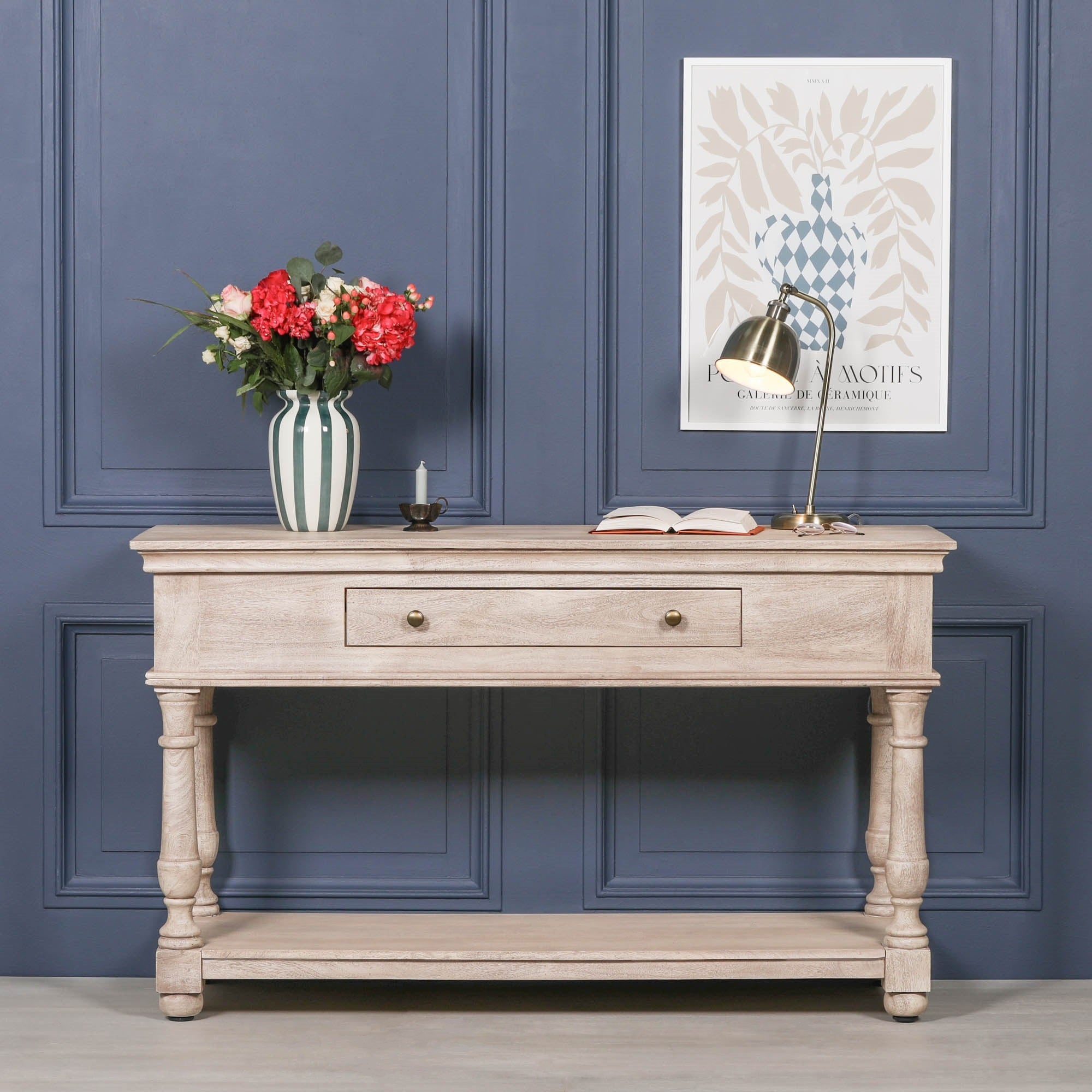 Camille acacia wood console table with drawer and lower shelf styled with flowers and lamp against a blue panelled wall