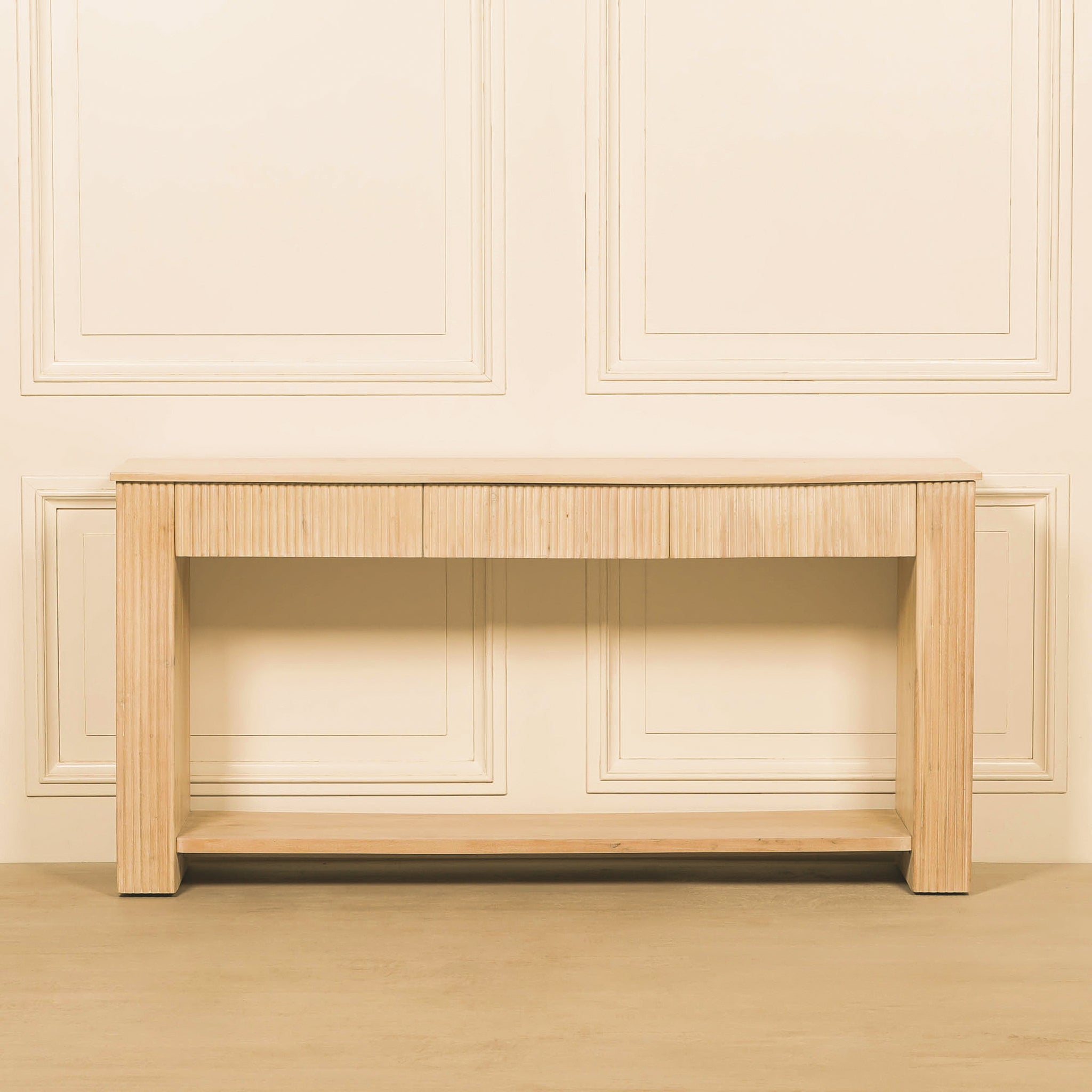 Front view of the Camille bleached wood console table with three fluted drawer fronts, substantial straight legs and a full-width lower shelf against a traditional panelled wall.