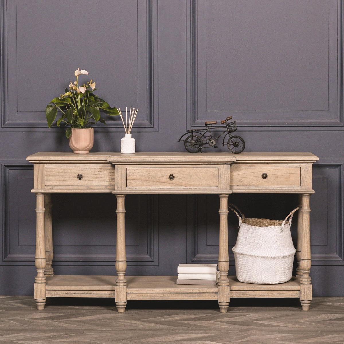 Camille Breakfront white cedar wood console table with three drawers in a rustic finish, styled against a dark panelled wall with decorative accessories, plant, reed diffuser and lower storage shelf, creating an elegant hallway or living room display.