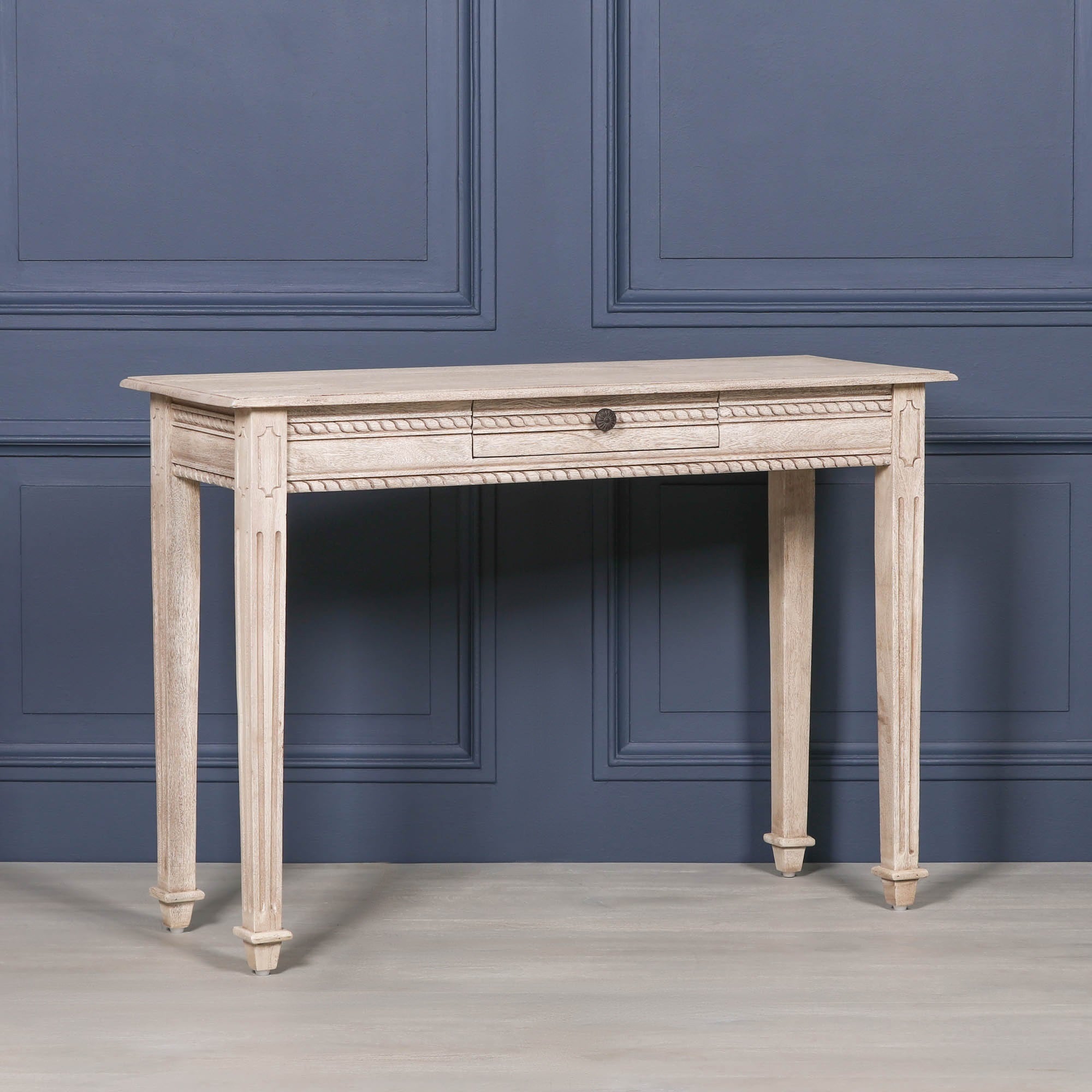 Angled view of Camille carved acacia wood dressing table with drawer against a dark blue panelled wall