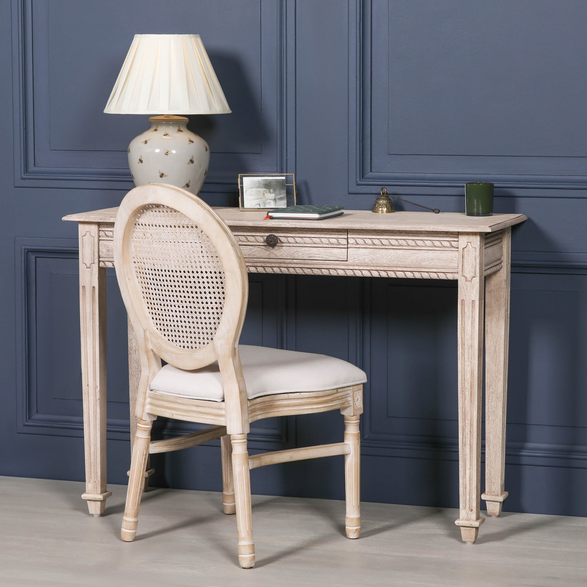 Camille carved acacia wood dressing table styled with rattan chair and lamp against a dark blue panelled wall