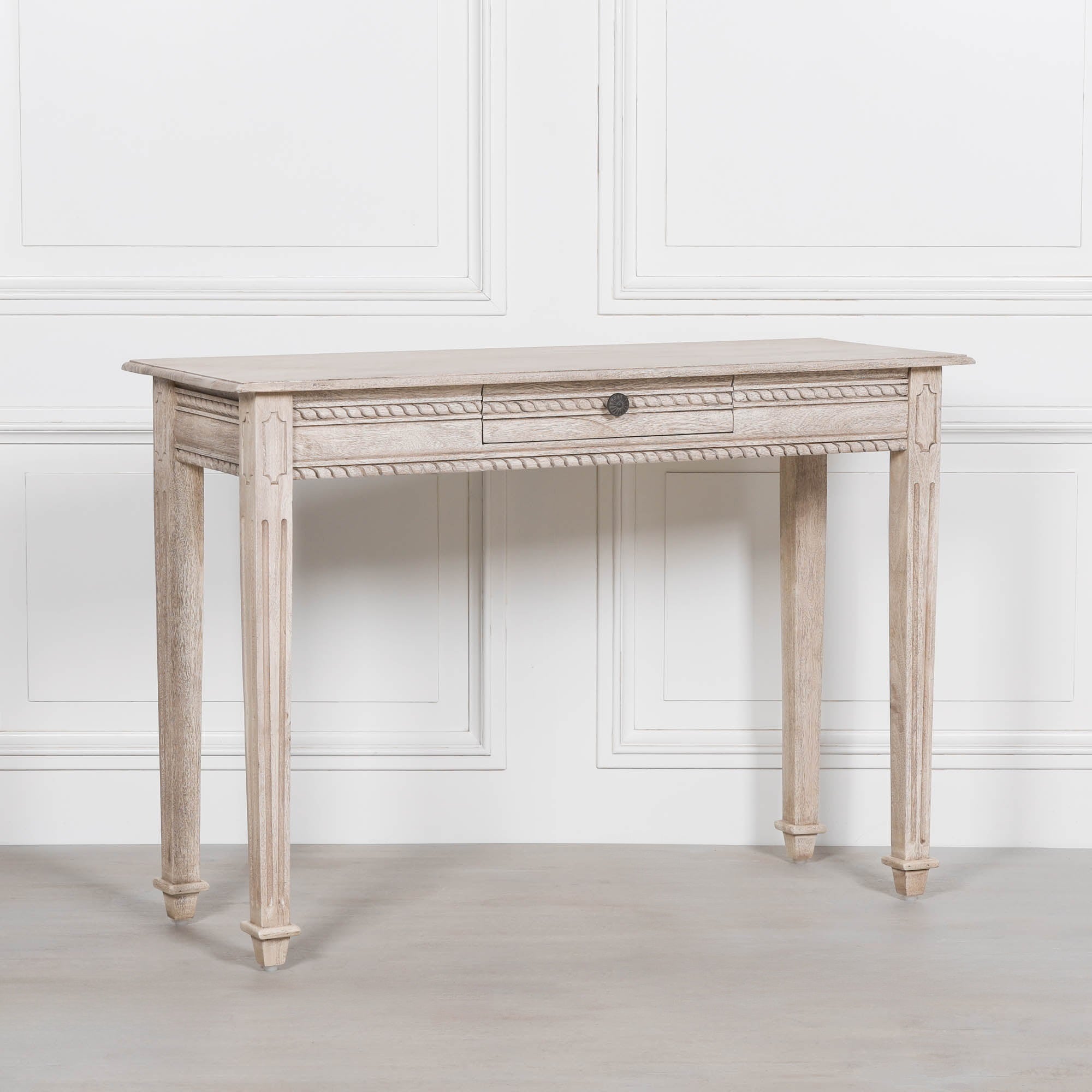 Angled view of Camille carved acacia wood dressing table with drawer against a white panelled wall
