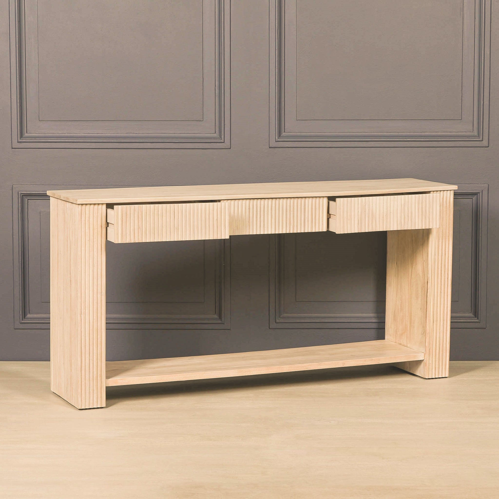 Angled view of the Camille fluted acacia wood console table against a charcoal panelled wall, featuring three storage drawers, textured detailing and an open lower shelf.