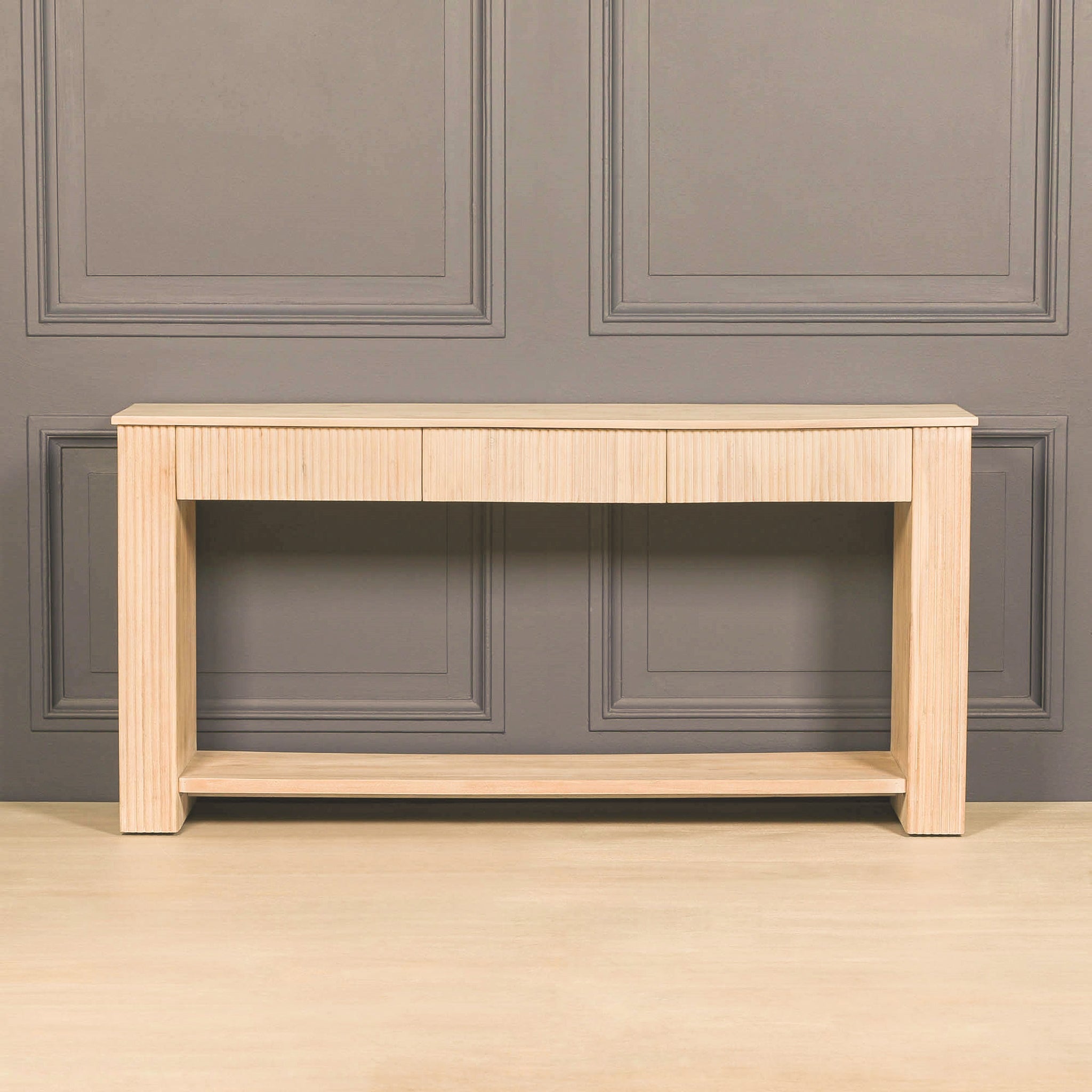 Camille fluted acacia wood console table shown against a dark panelled wall, with three discreet drawers, vertically textured detailing and a long open lower storage shelf.