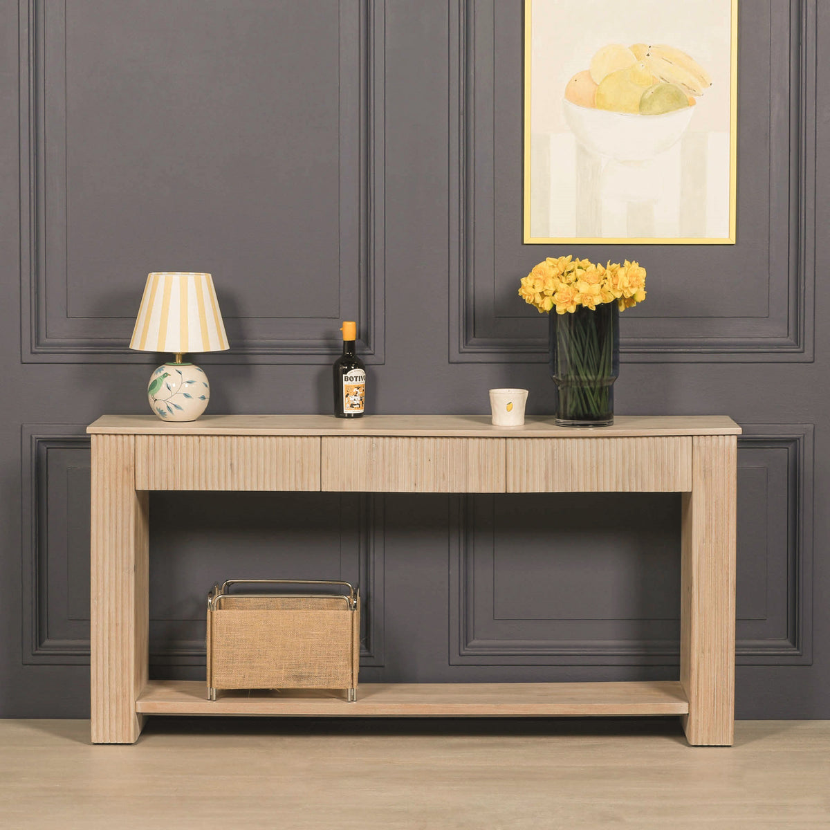 Camille fluted acacia wood console table styled against a dark panelled wall, featuring three drawers, a long lower display shelf and a light bleached washed wood finish.