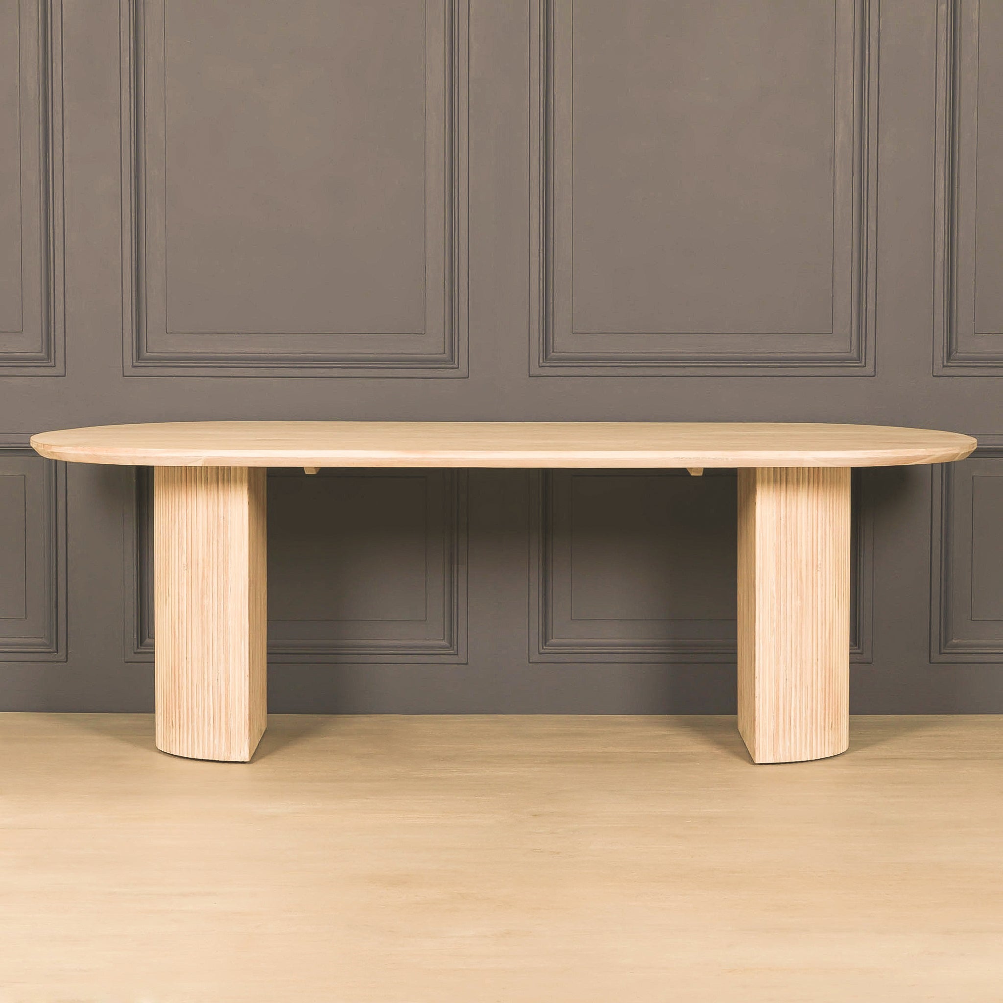Camille Fluted Acacia Wood Oval 6 Seater Dining Table in bleached washed acacia wood against a grey panelled wall, showcasing the elegant oval tabletop, fluted pedestal legs and natural wood grain finish.