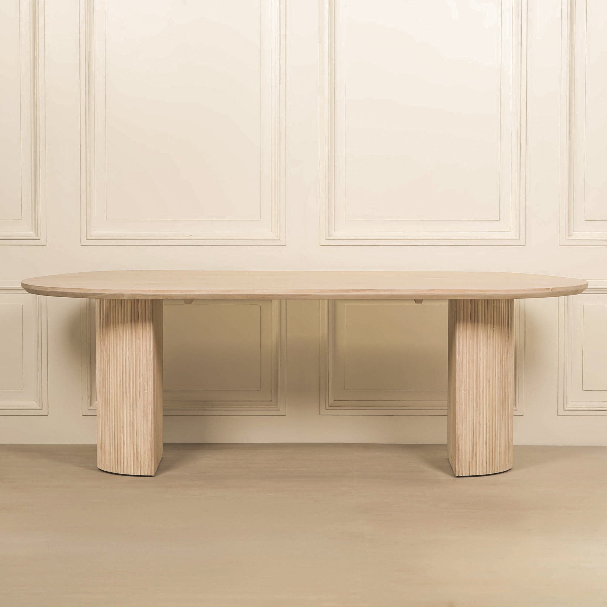 Camille Fluted Acacia Wood Oval 6 Seater Dining Table with a bleached washed wood finish displayed against a neutral panelled backdrop, highlighting its oval silhouette, contemporary fluted pedestal base and solid acacia wood construction.