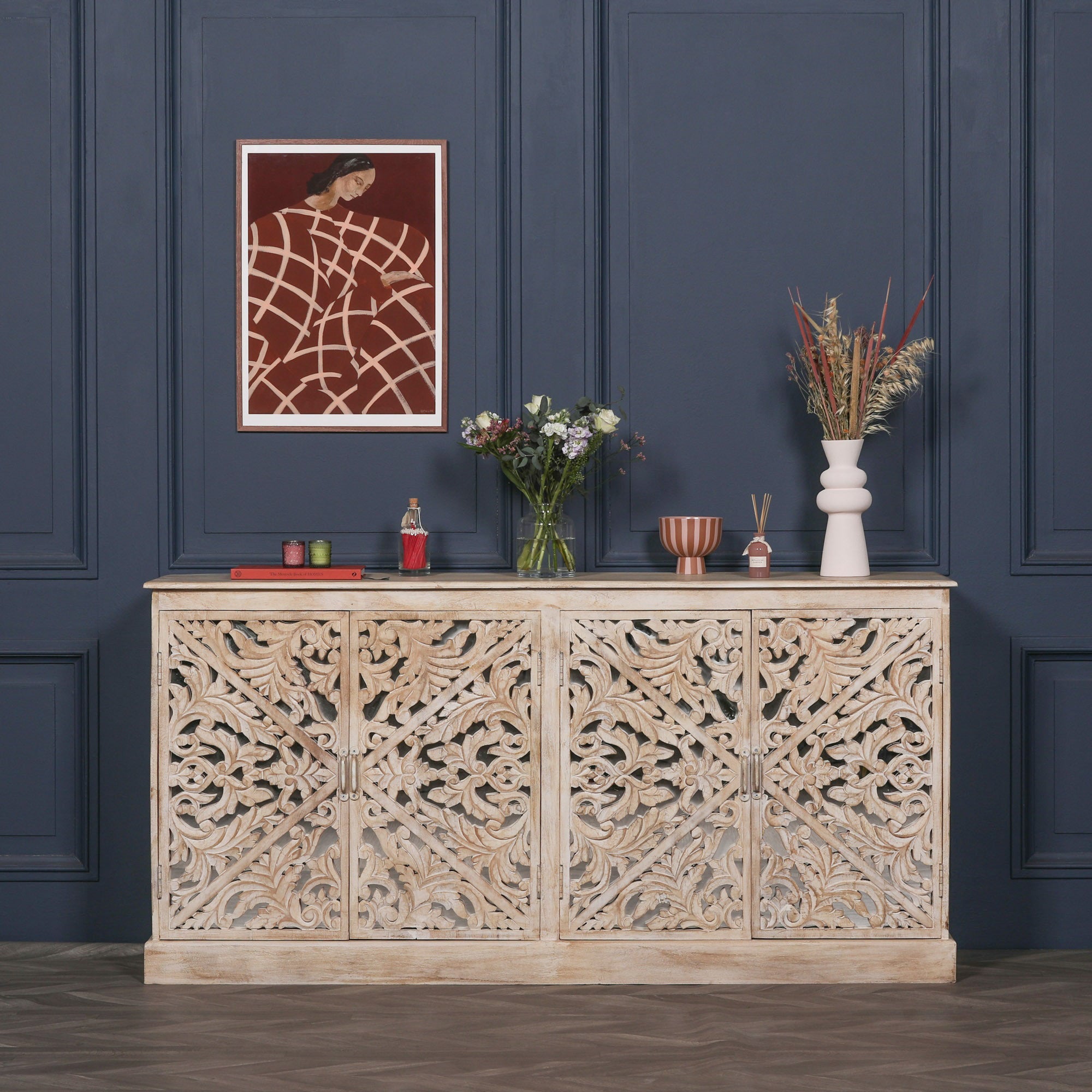 Camille Four Door Carved Mirrored Sideboard in bleached mango wood styled in a luxury living room with decorative accessories, handcrafted carved doors, mirrored panels and spacious concealed storage.