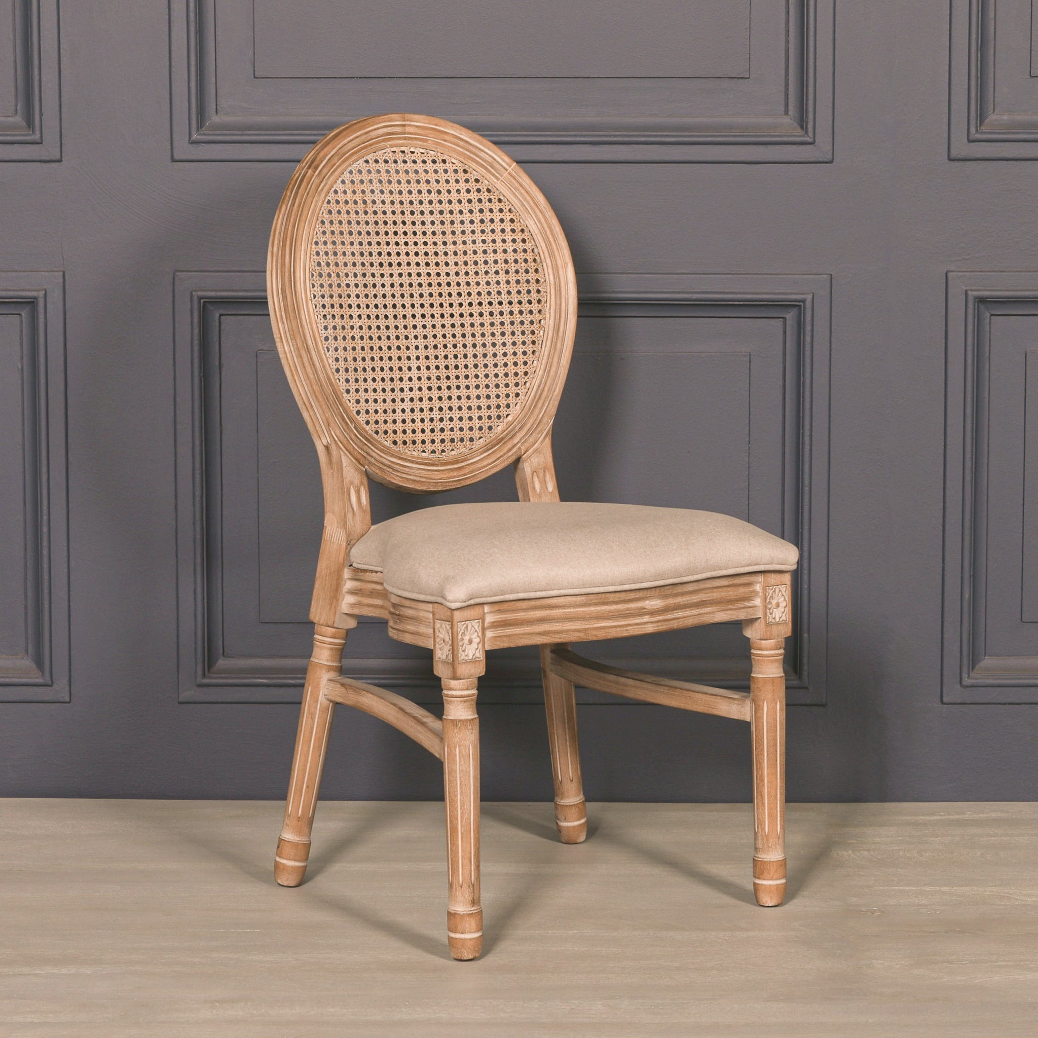 Three-quarter front view of the Camille French Louis Linen Upholstered Dining Chair featuring a carved natural wooden frame, woven cane backrest and soft linen seat against a charcoal panelled backdrop.