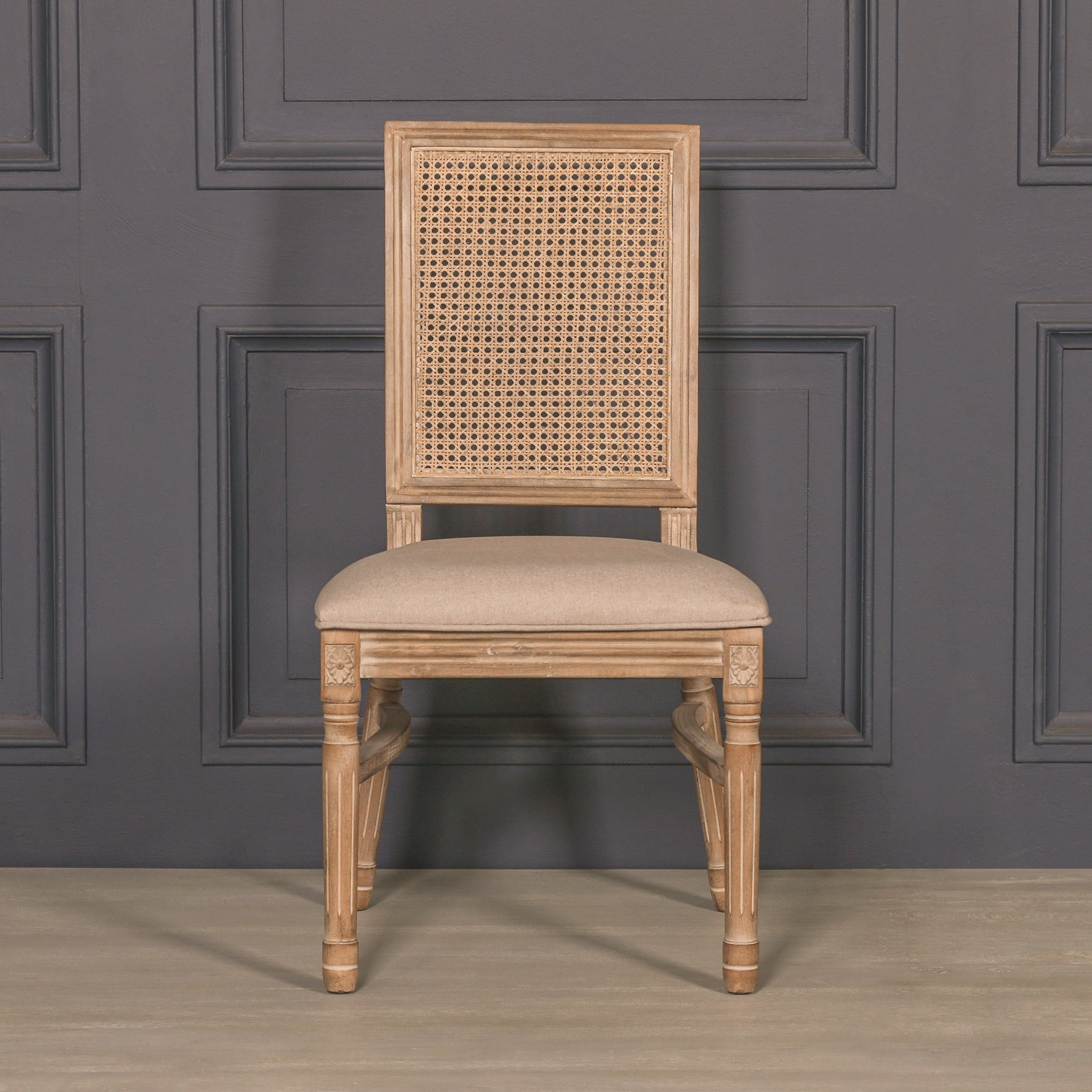 Front view of the Camille French Louis Rattan Back Dining Chair with natural rattan back, linen upholstered seat and carved wooden frame against a dark panelled wall.